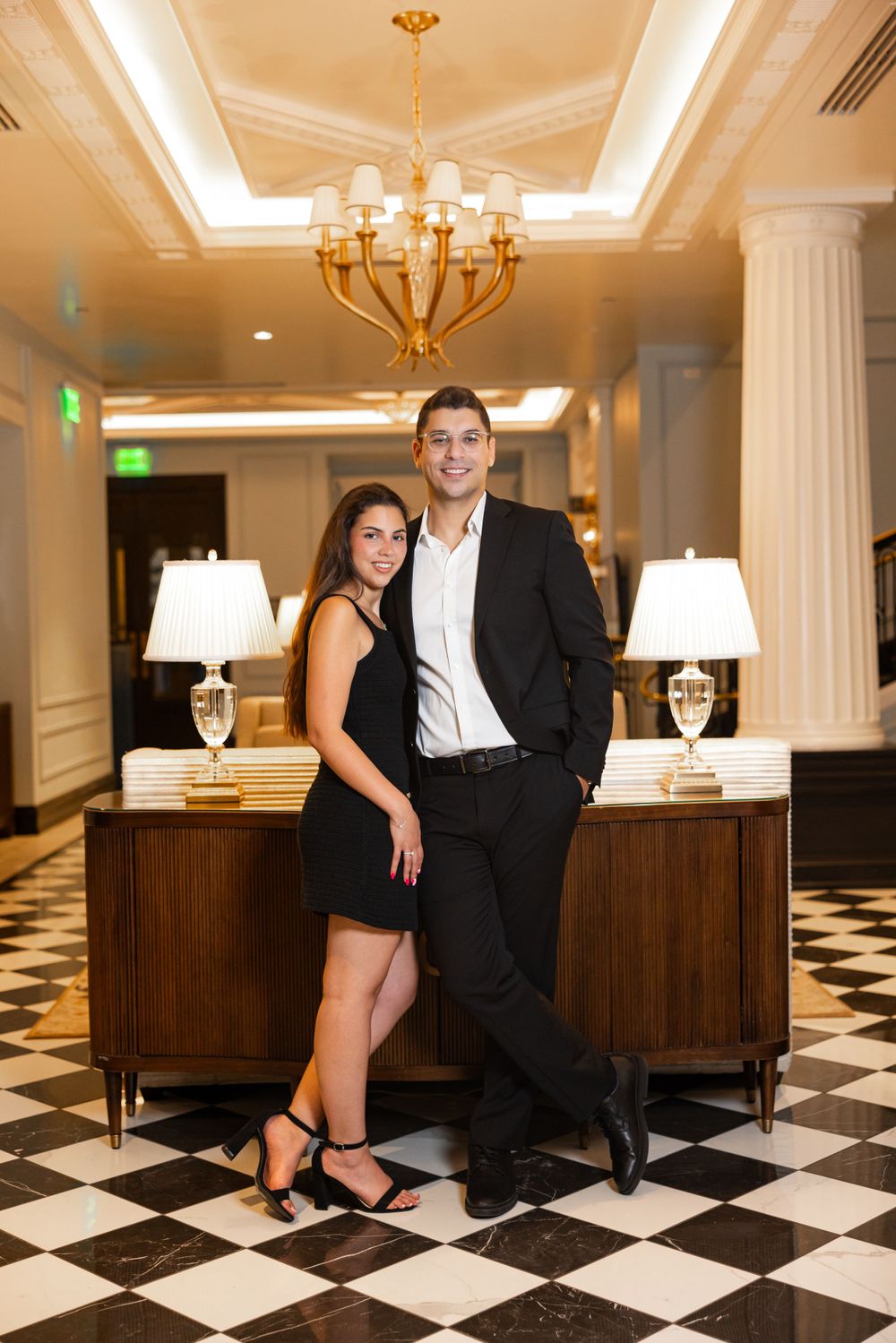 Elegant couple posing against luxurious hotel interior with chandelier and checkered floor.