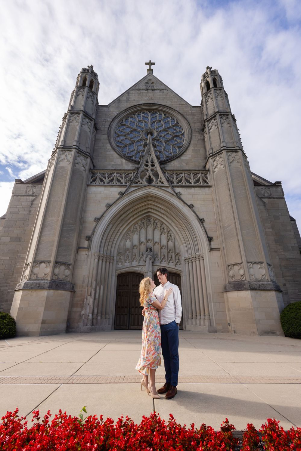 A couple embraces in front of a Gothic cathedral with red flowers blooming in the foreground.