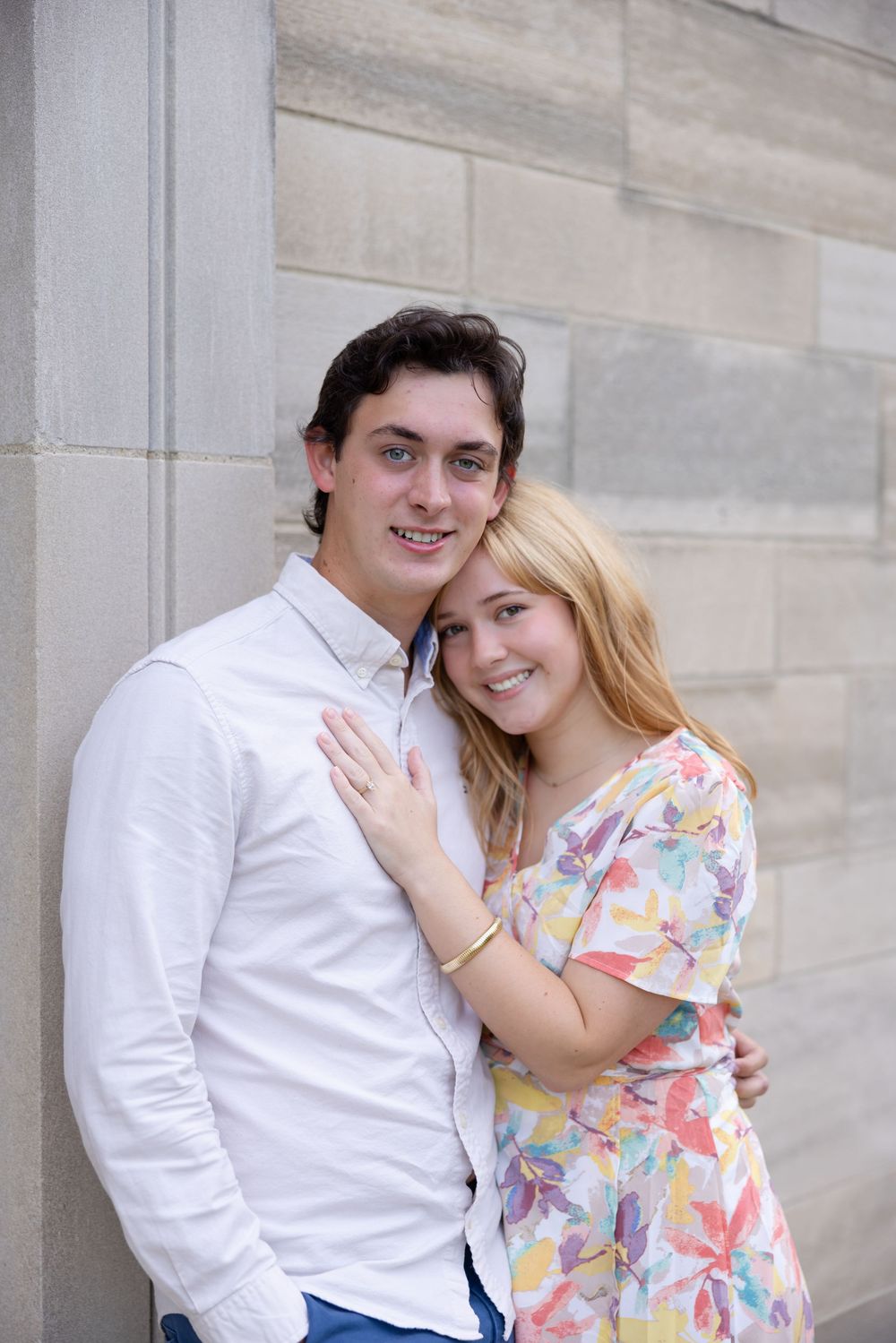 A couple poses together against a stone wall, with one wearing a white shirt and the other in a floral dress.