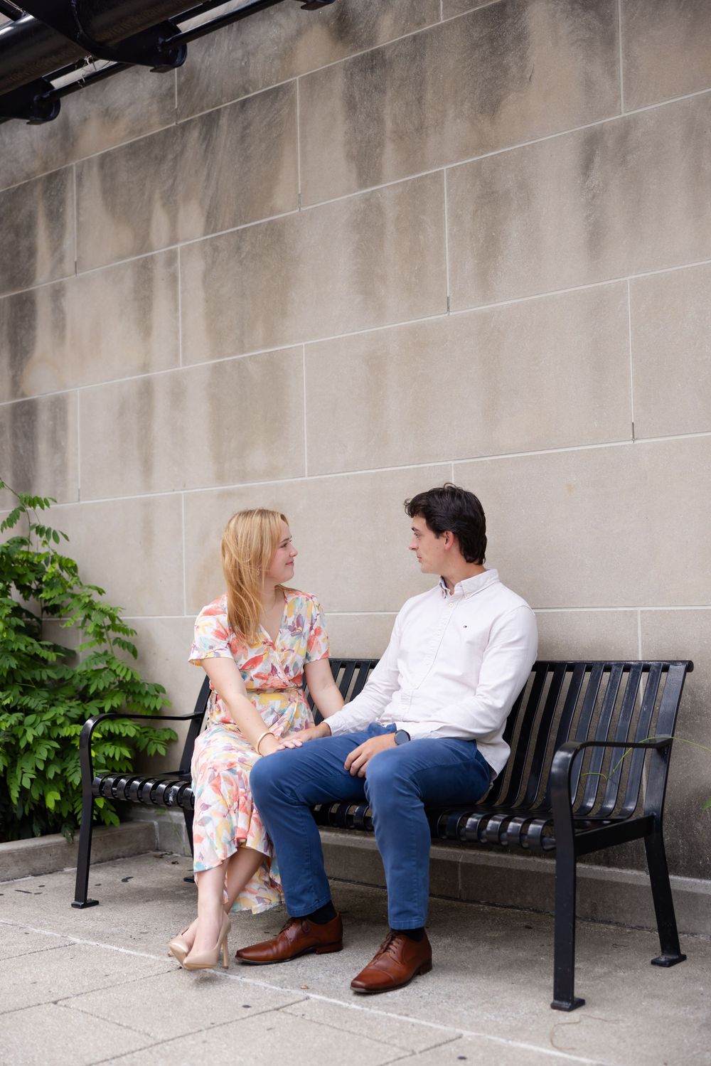 A couple sits together on a black metal bench against a concrete wall with greenery on a summer day.