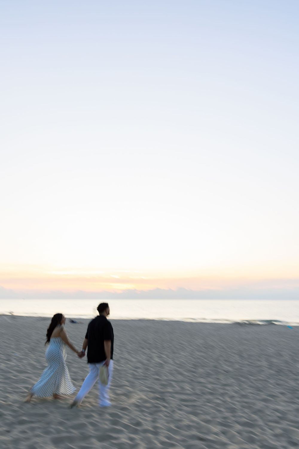 At Indiana Dunes, Two figures in white outfits walk together along a sandy beach at sunset creating a romantic silhouette.