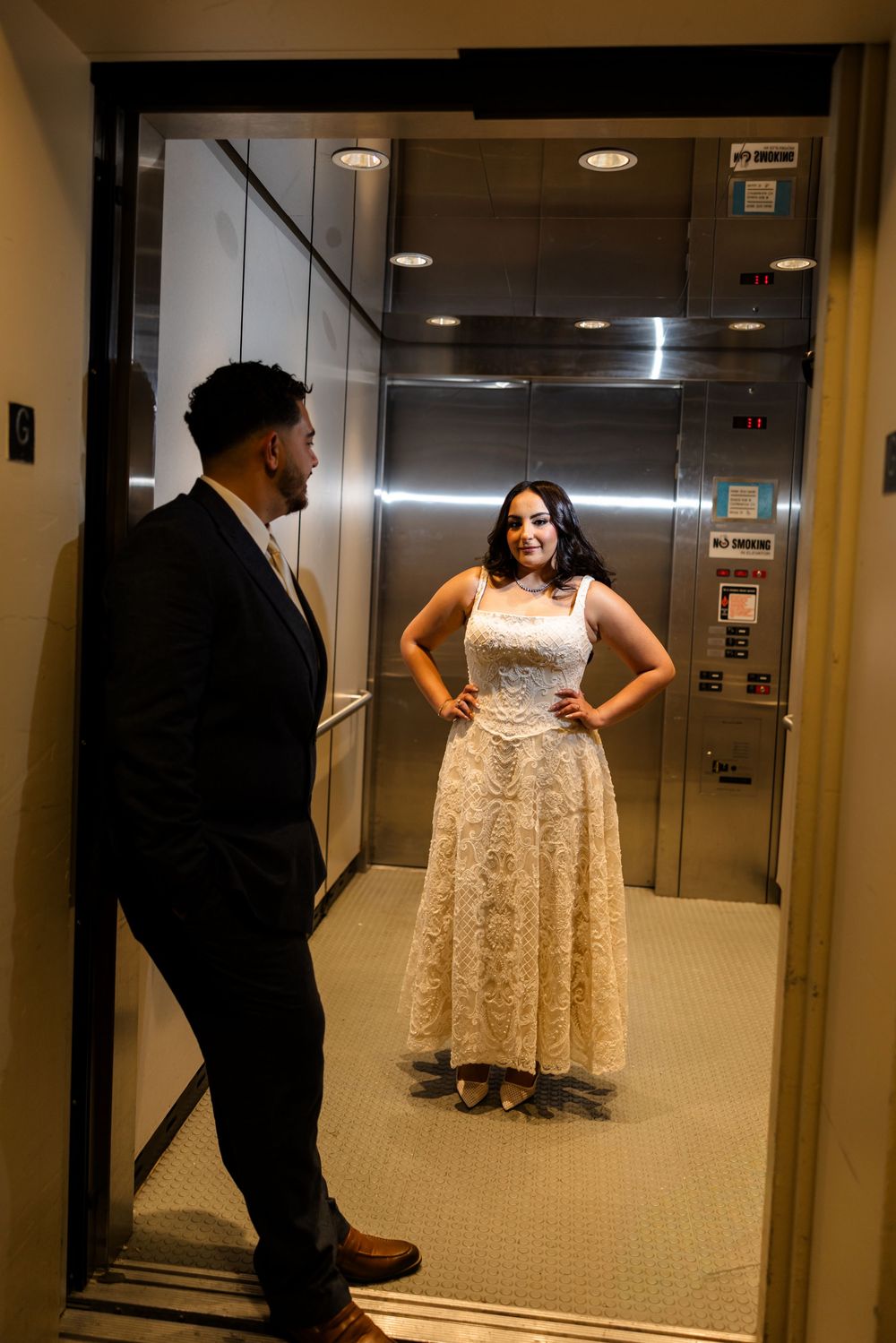 Formal couple poses playfully in a mirrored elevator lobby area.