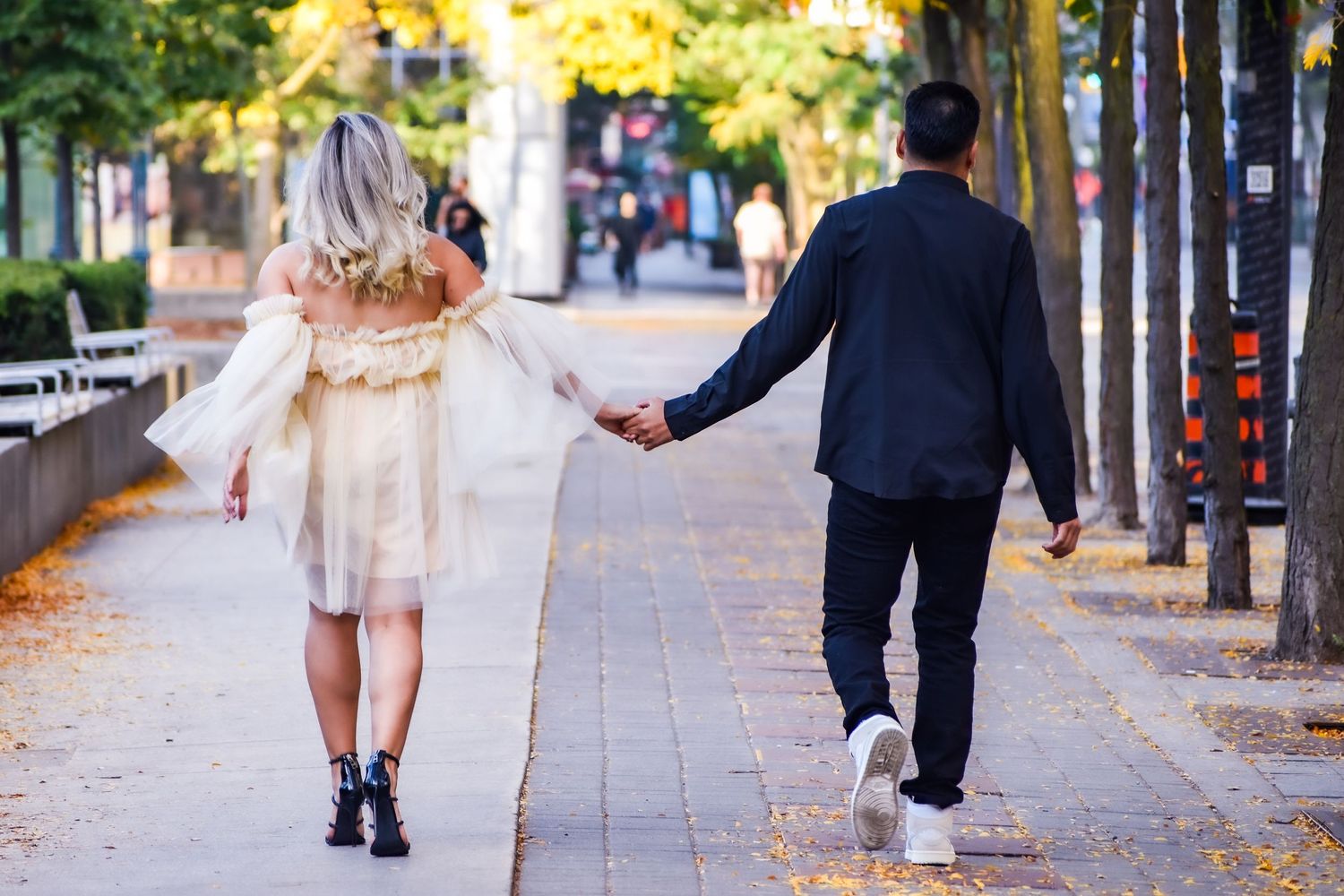 Couple holding hands walking down a tree-lined urban sidewalk in autumn. Toronto GTA