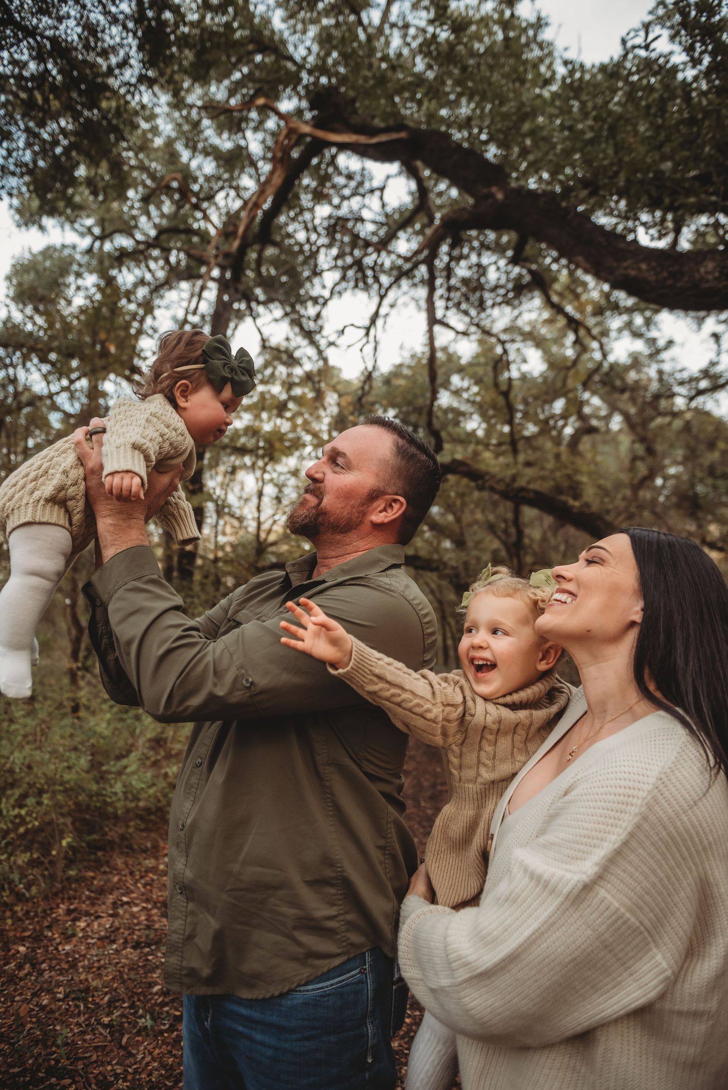 Family enjoying playful moment together outdoors under trees during autumn photoshoot.