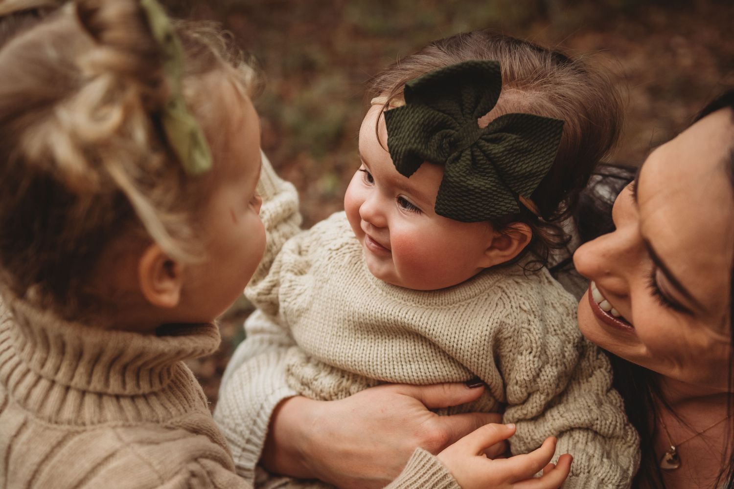 A baby in a cream knit sweater and dark bow headband shares a tender moment with family members.