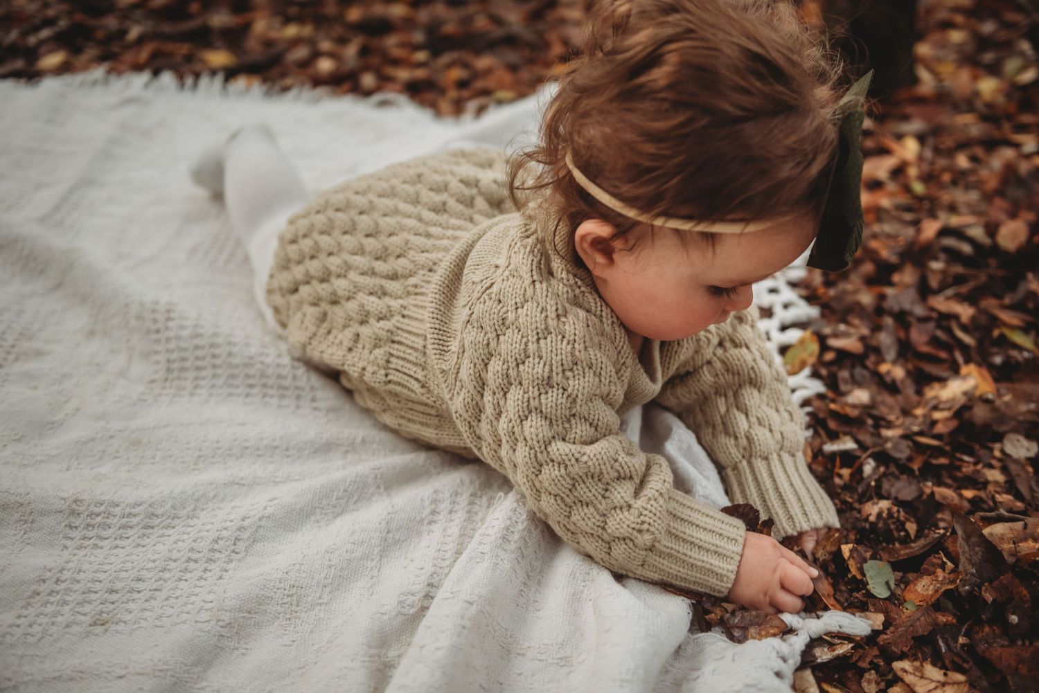 Someone in a cozy knit sweater lies on a white blanket surrounded by fallen autumn leaves.