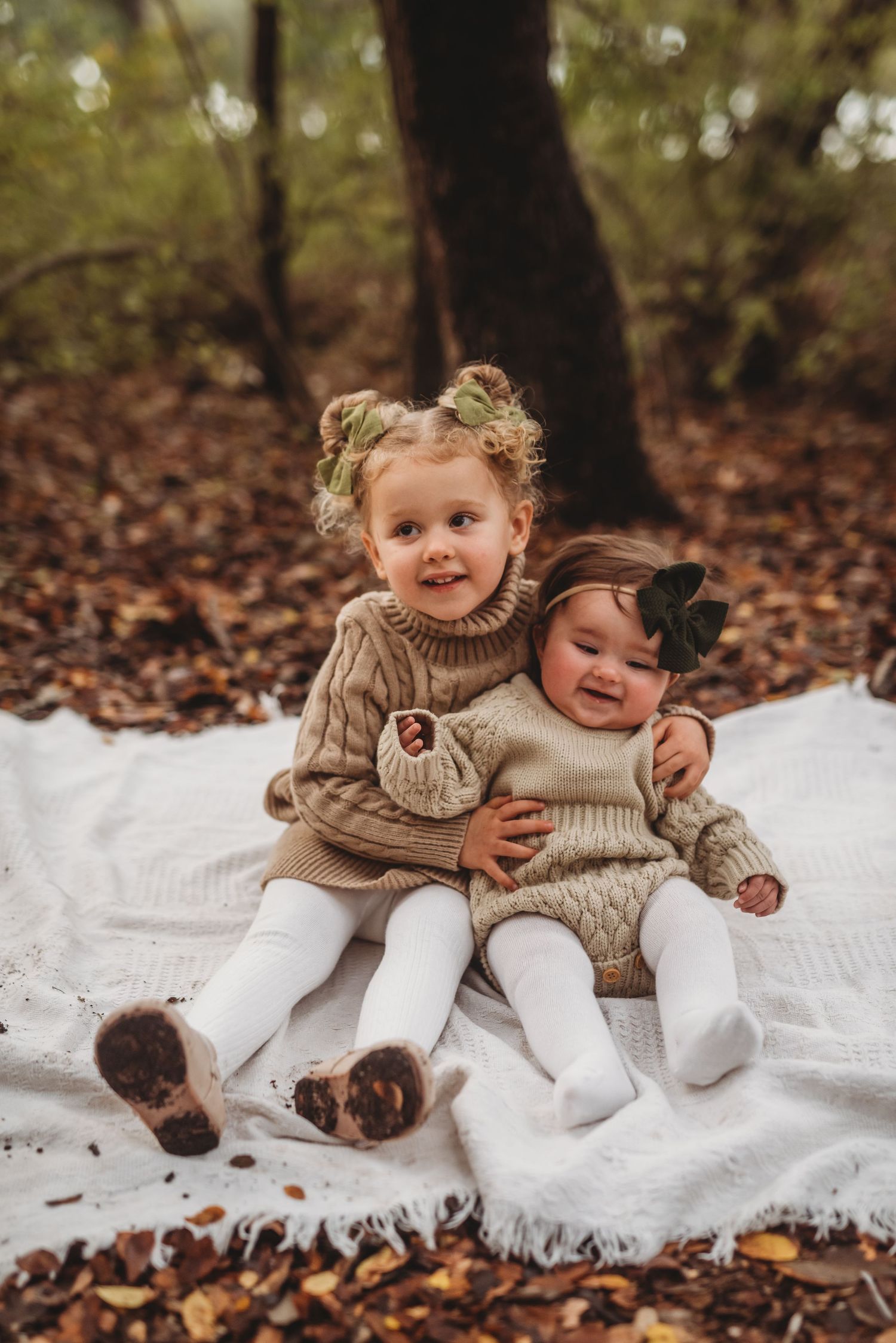 Two young children in matching knit sweaters sitting together on a white blanket surrounded by autumn leaves.