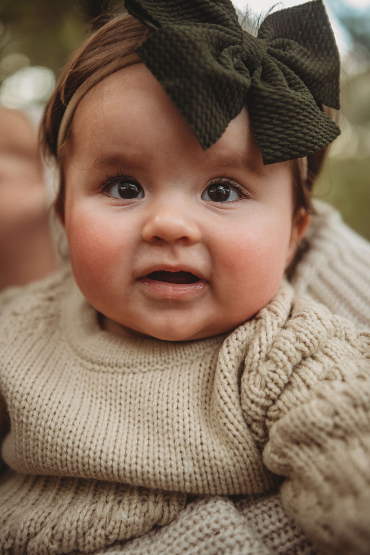 Baby wearing cozy beige knit sweater and olive green bow headband against blurred background.