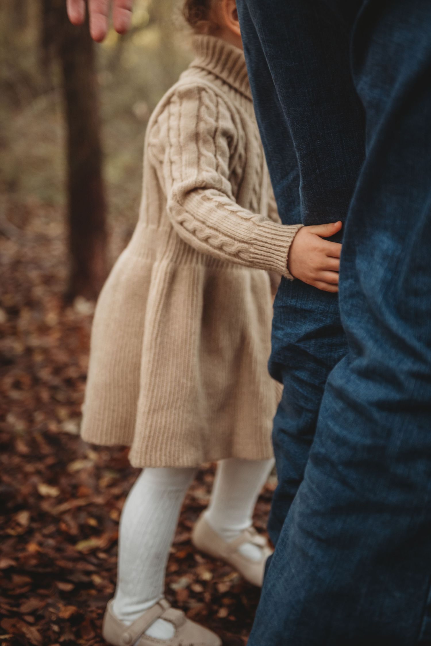 Close-up of child in beige knit dress holding parent's leg among fallen leaves.
