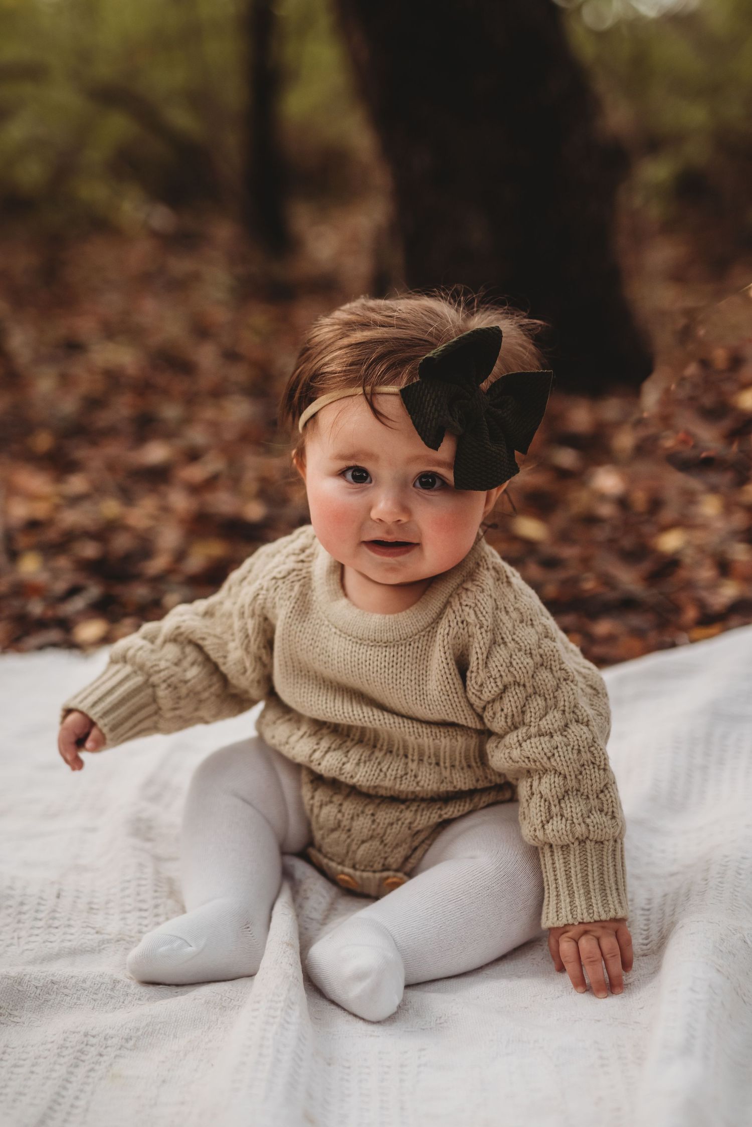 Baby wearing knit beige sweater and white tights sits on blanket in autumn forest setting.