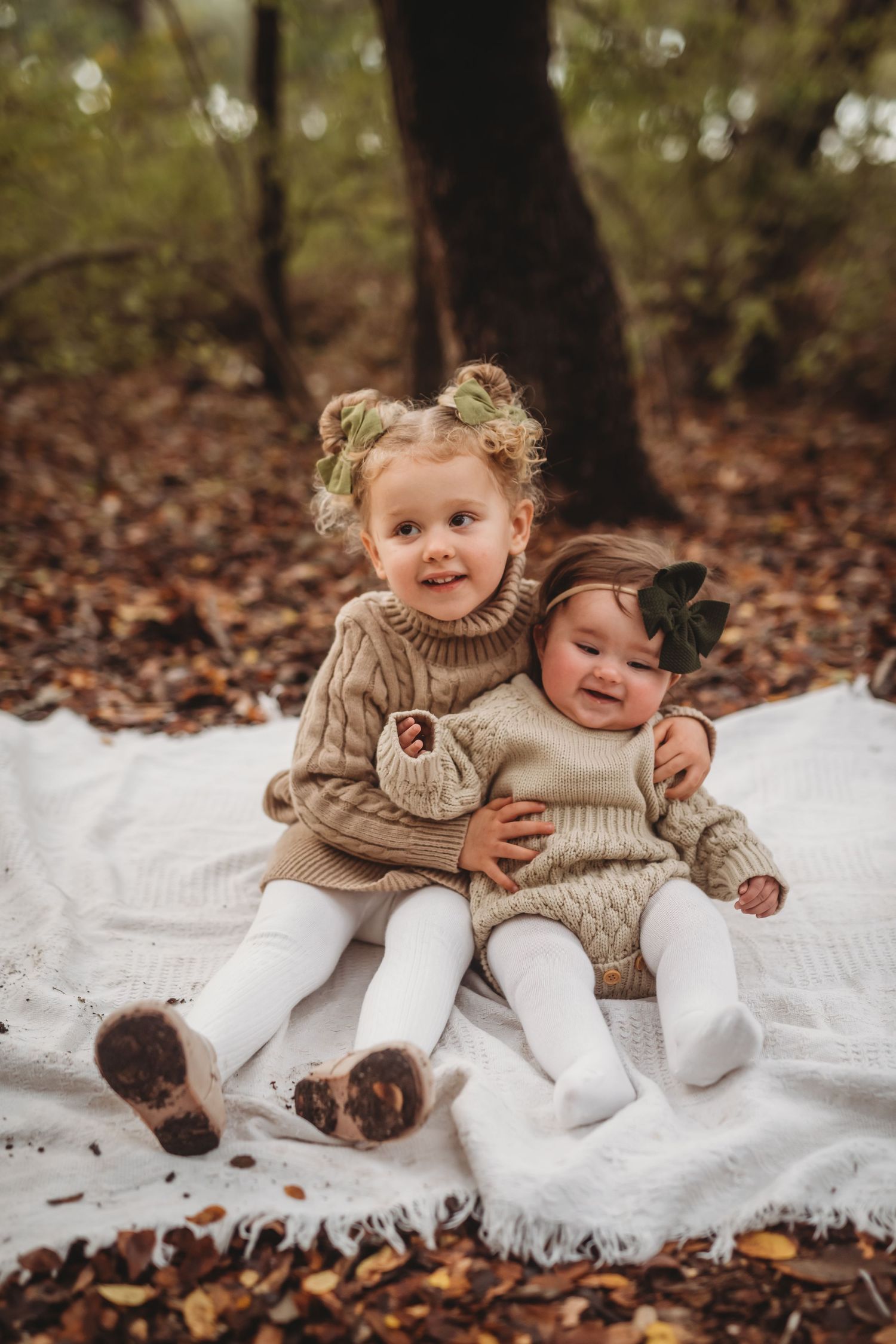 Two children in cozy knit sweaters sit together on a white blanket surrounded by fallen leaves in the woods.