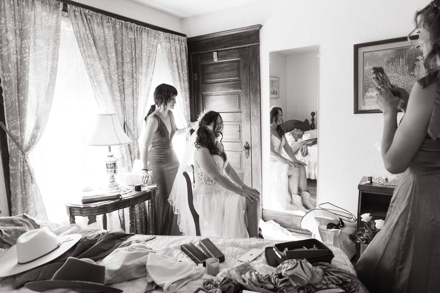 A black and white photo of wedding preparation in a bedroom with clothing and people getting ready.