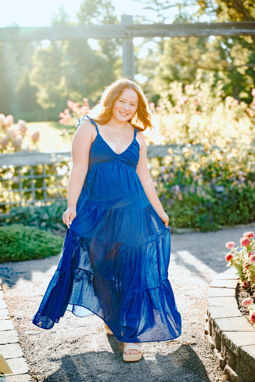 A flowing royal blue dress is showcased in a sunlit garden with colorful flowers and stone pathways.