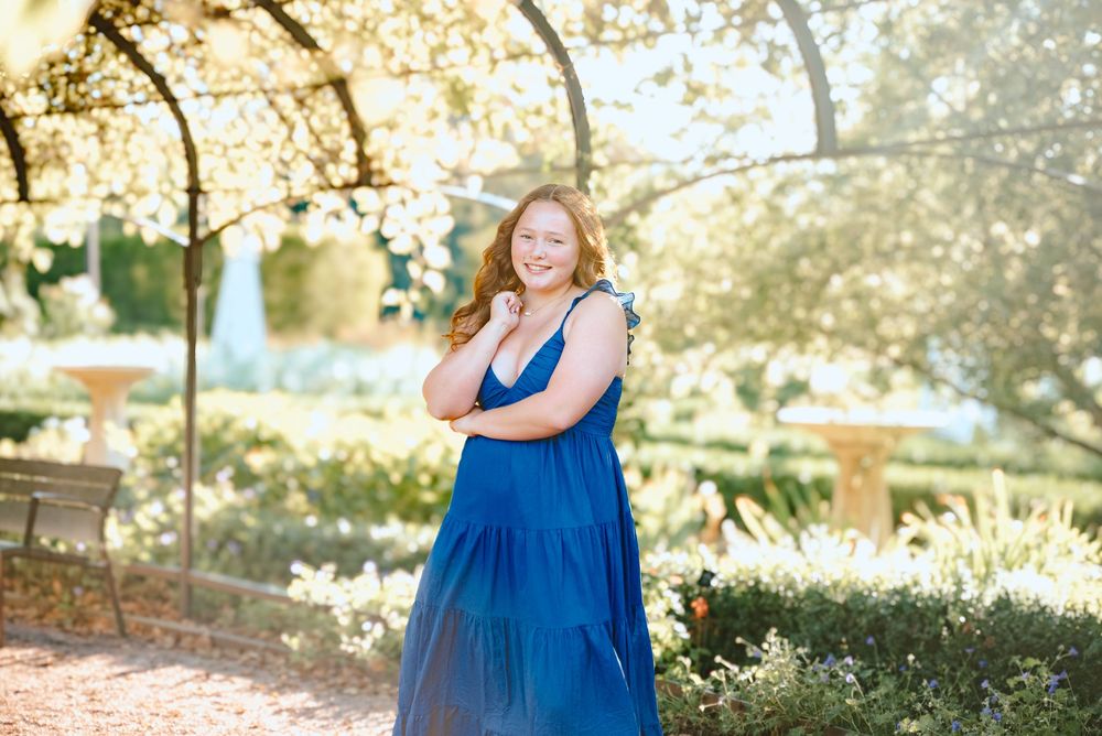 A sunlit garden path with an iron arbor framing someone in a flowing tiered blue maxi dress.