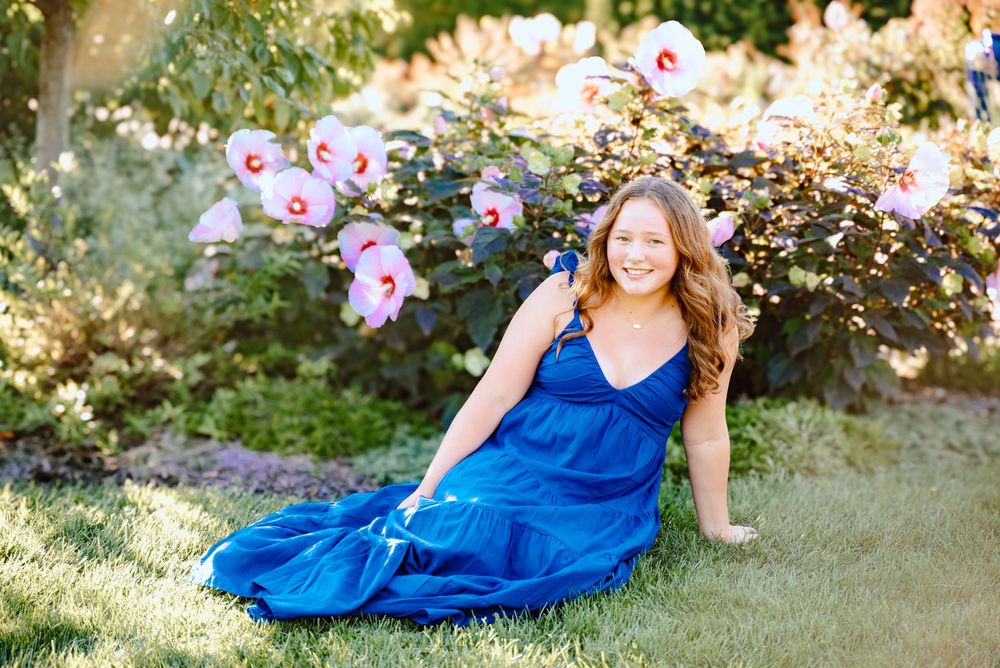 Person in a flowing royal blue dress poses playfully in a sunlit garden setting.