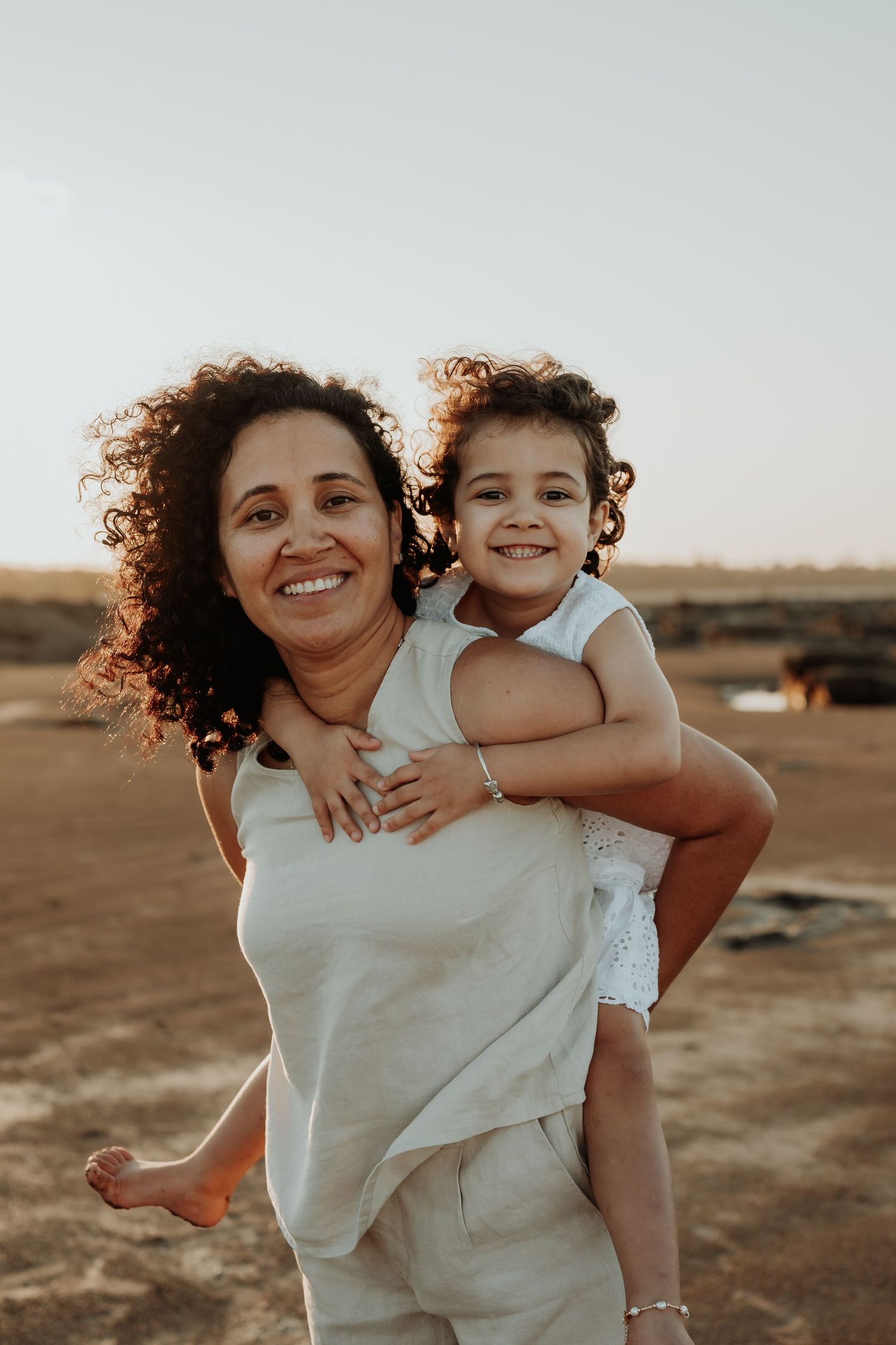 A heartwarming piggyback ride moment captured at sunset on a Gladstone beach with natural curly hair flowing in the golden light.