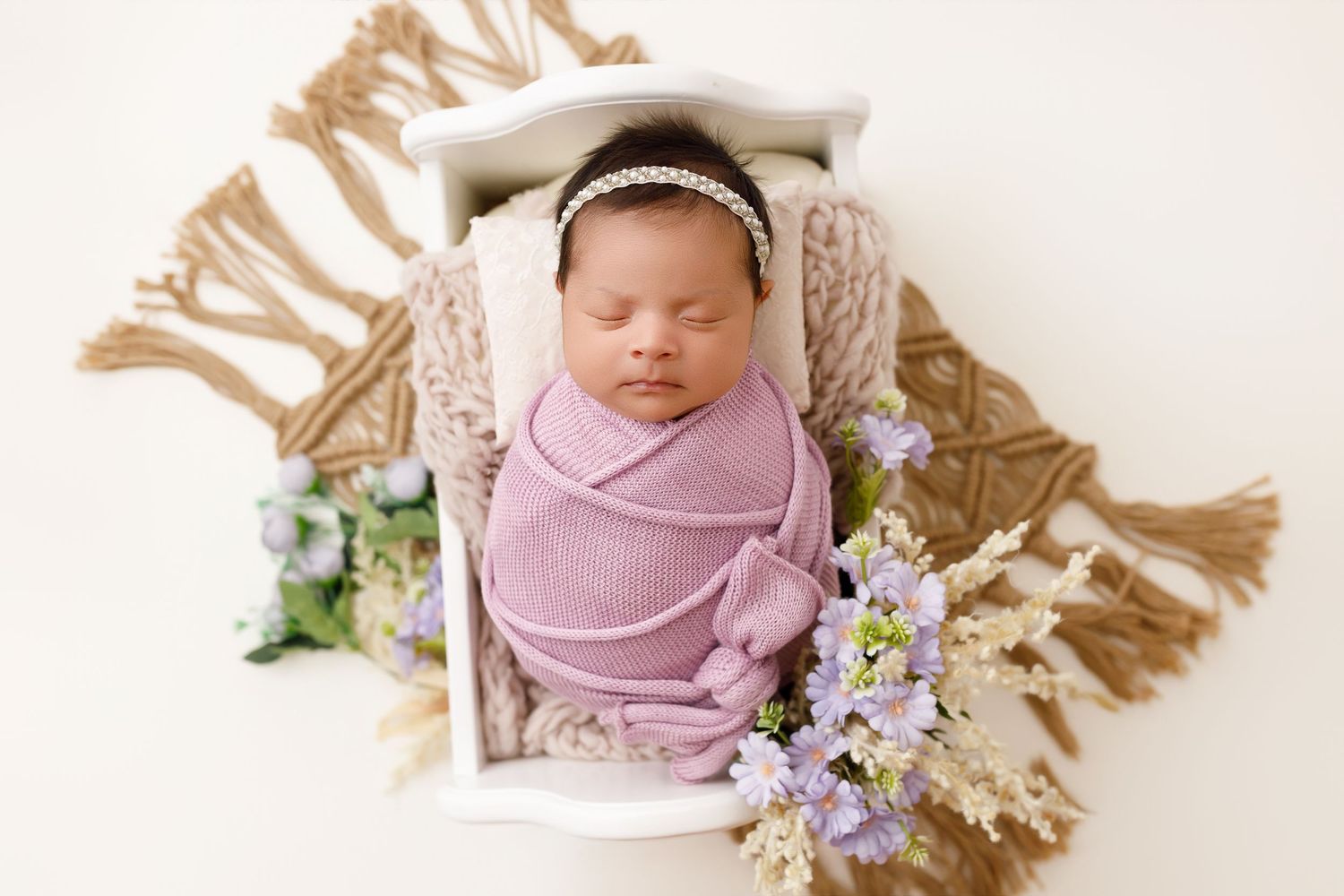 Sleeping newborn wrapped in purple swaddle with delicate flowers on white backdrop and woven mat.