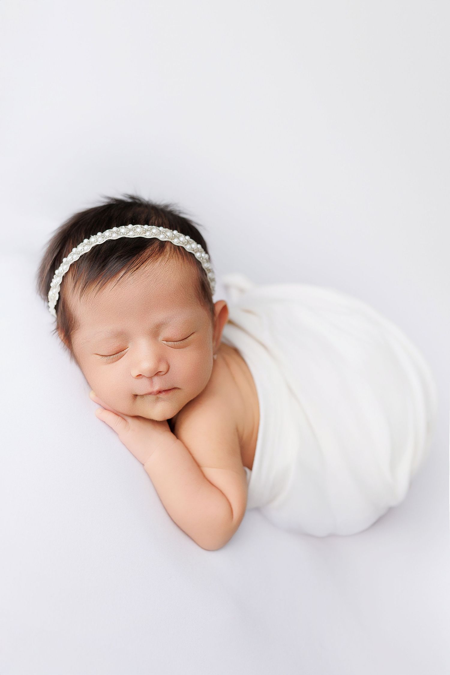A sleeping newborn baby wrapped in soft white fabric wearing a delicate headband in a minimalist studio setting.