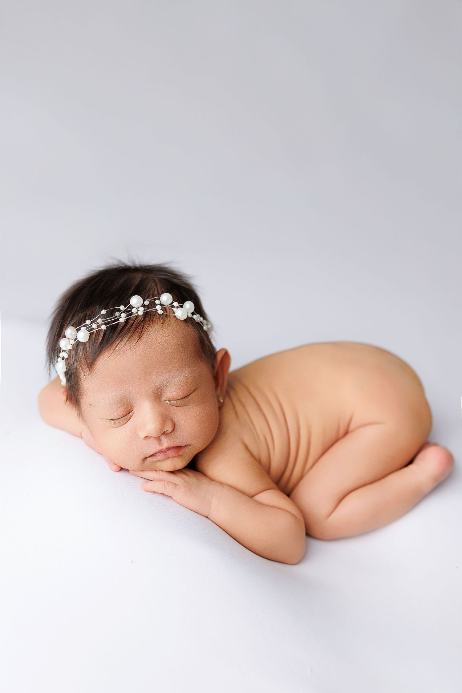 A sleeping newborn baby wearing a delicate floral headband lies peacefully on a white background in a minimalist portrait.