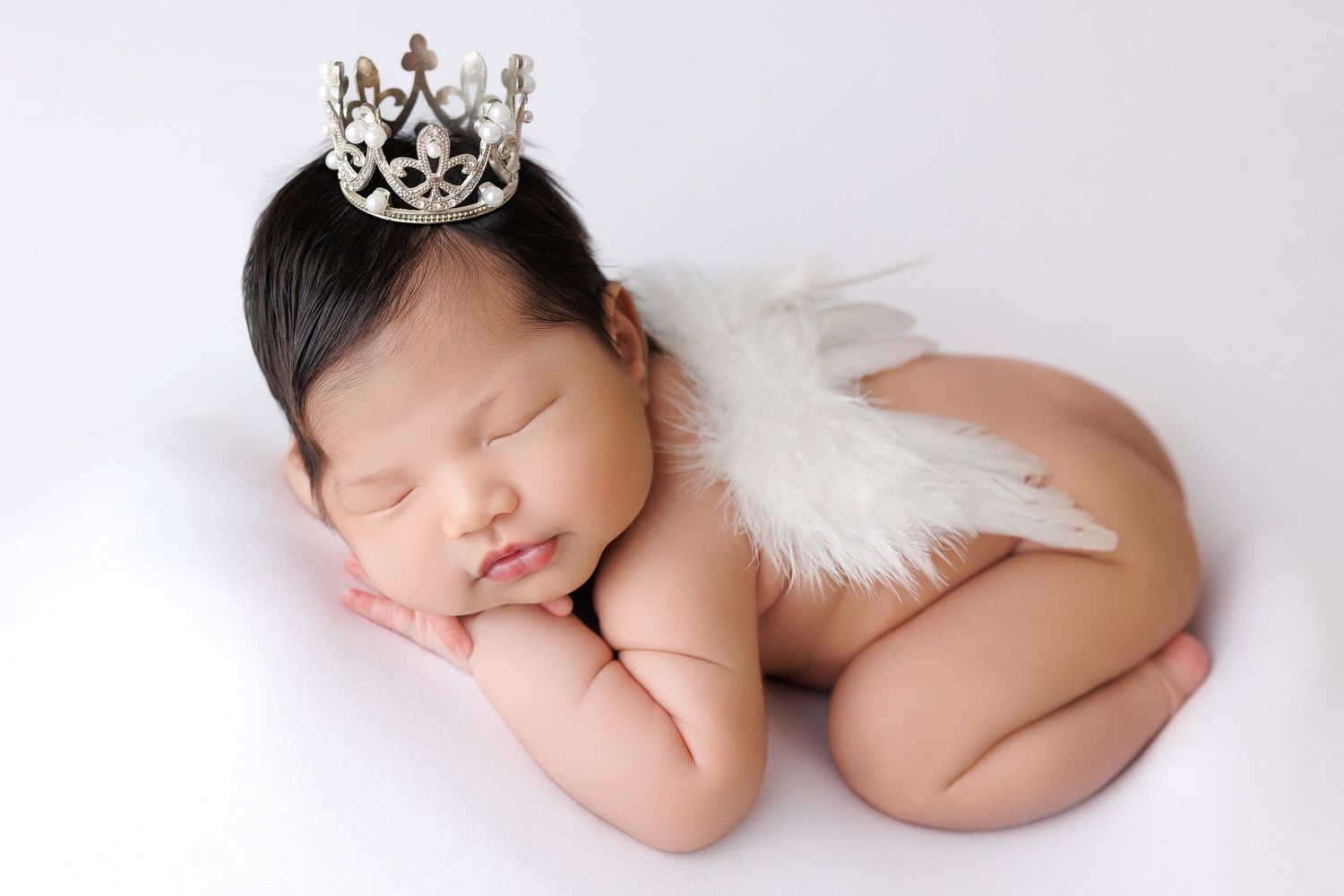 Sleeping newborn wearing small silver crown and angel wings lying peacefully on white background.