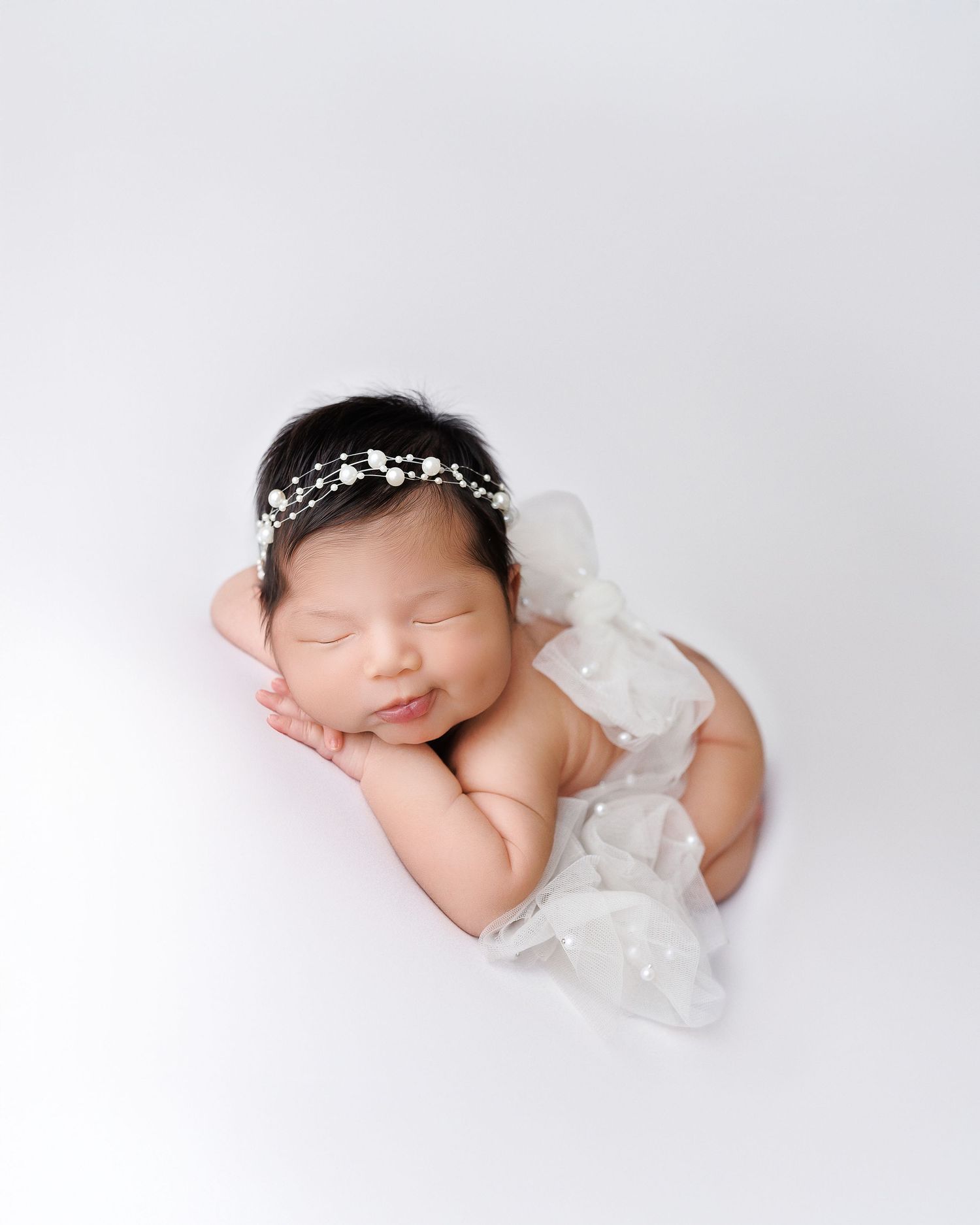 Sleeping newborn wearing delicate pearl headband and white wrap rests peacefully on white backdrop during photo session.