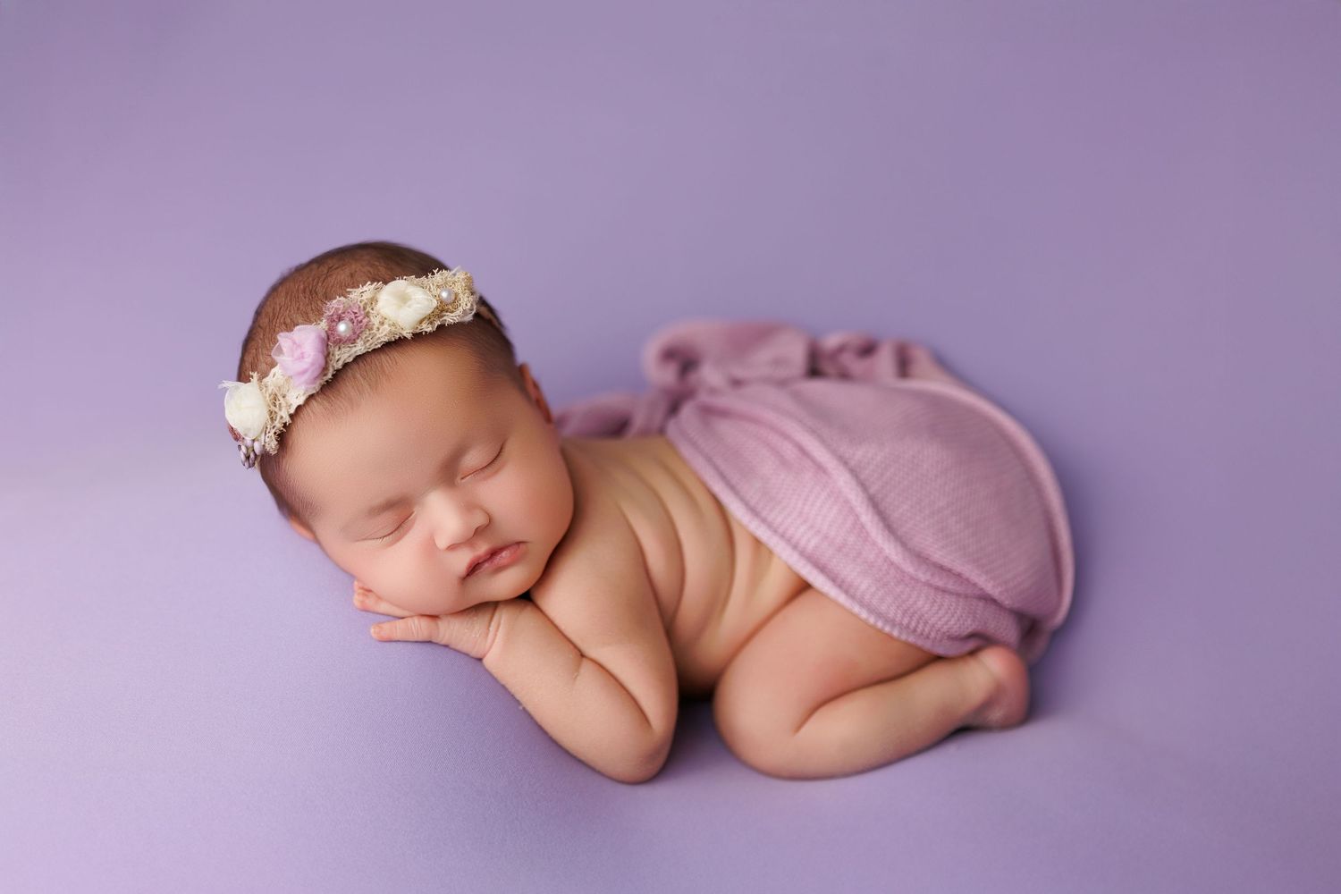 Sleeping newborn baby in purple hues curled up in fetal position wearing delicate floral headband.