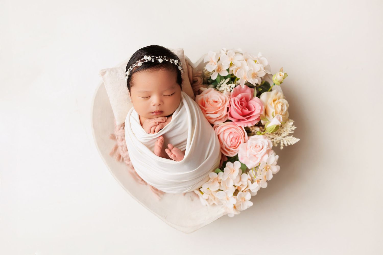 Four connected photos show a sleeping newborn baby swaddled in white fabric alongside soft pink and white flowers.