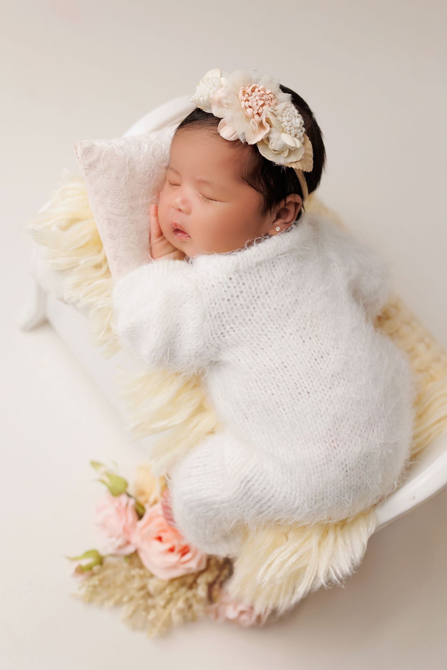 Newborn baby sleeping peacefully on cream fur blanket wrapped in white knit swaddle with floral headband and pink roses.