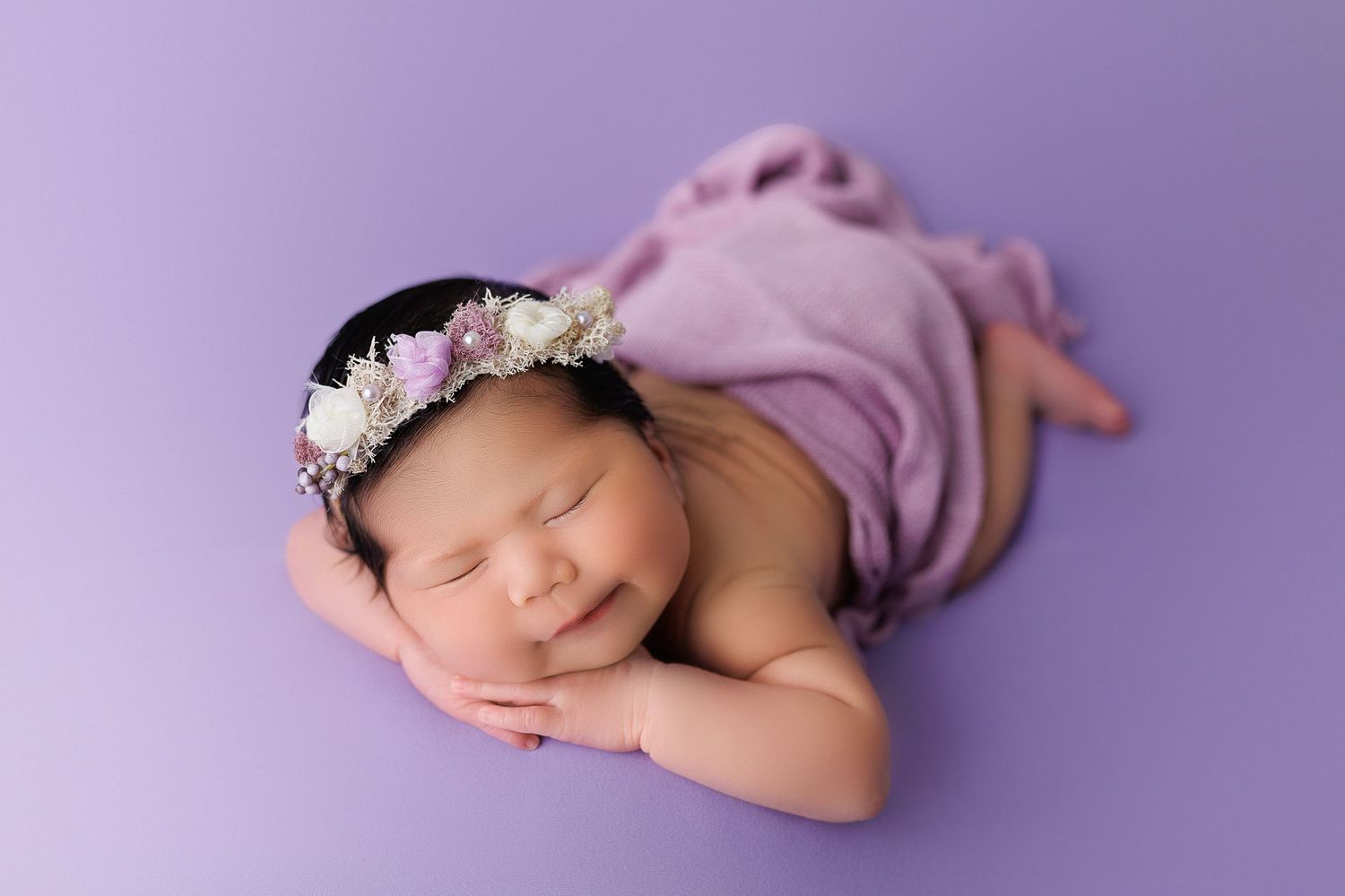 Sleeping newborn wearing floral crown on purple backdrop.