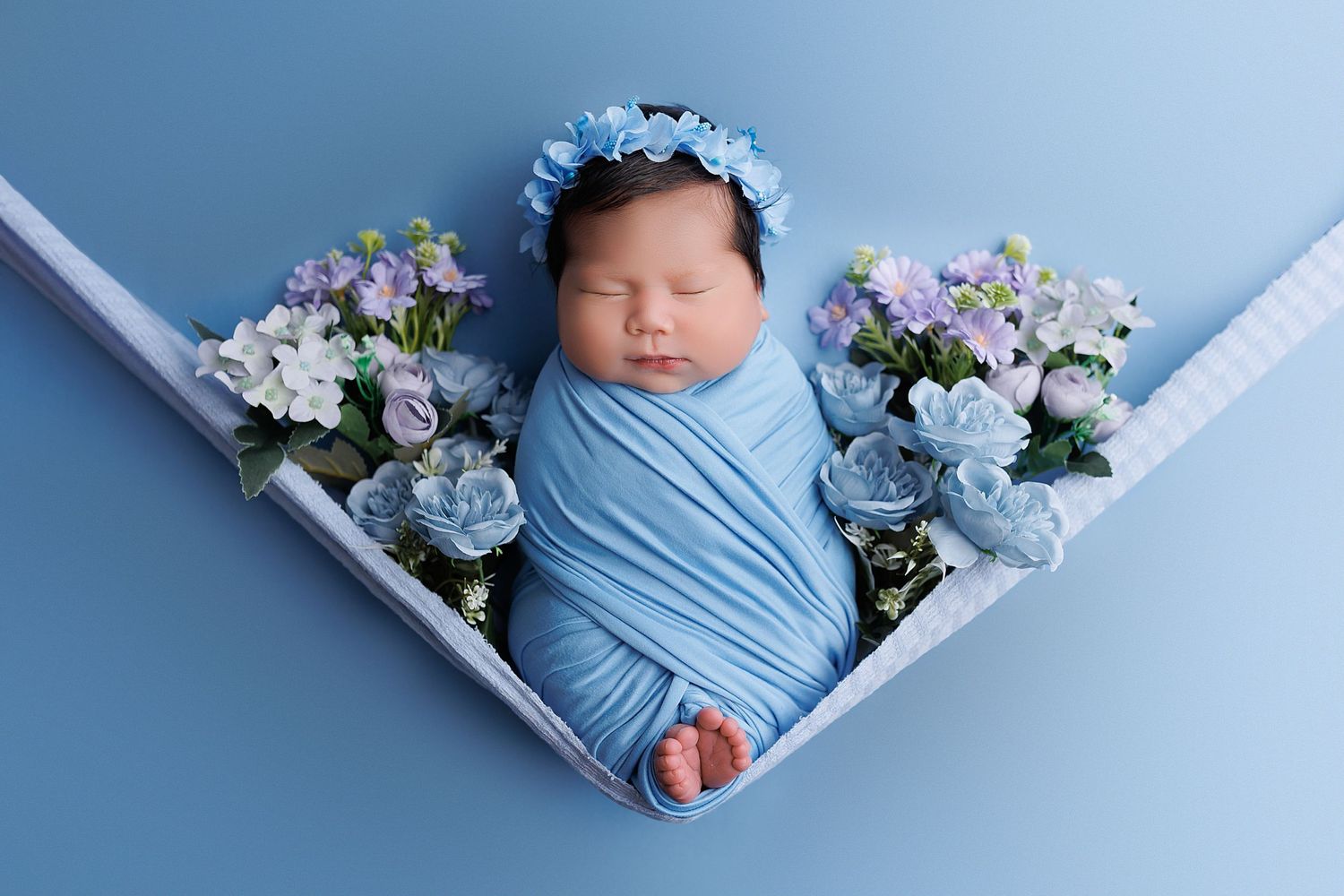 Newborn wrapped in blue swaddle with matching floral crown and hydrangea decorations.