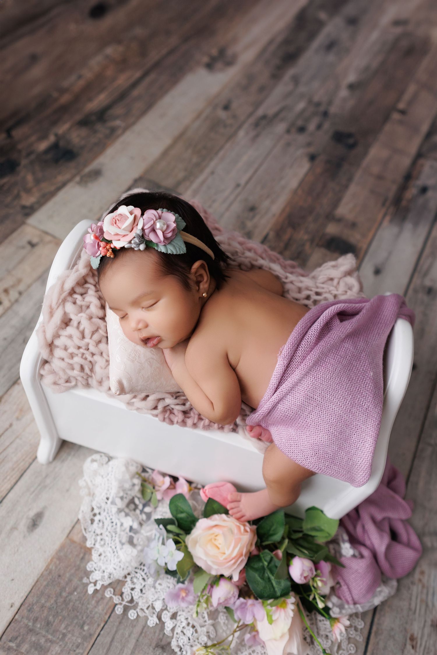 Sleeping newborn on white bed with pink floral headband and roses on wooden floor.