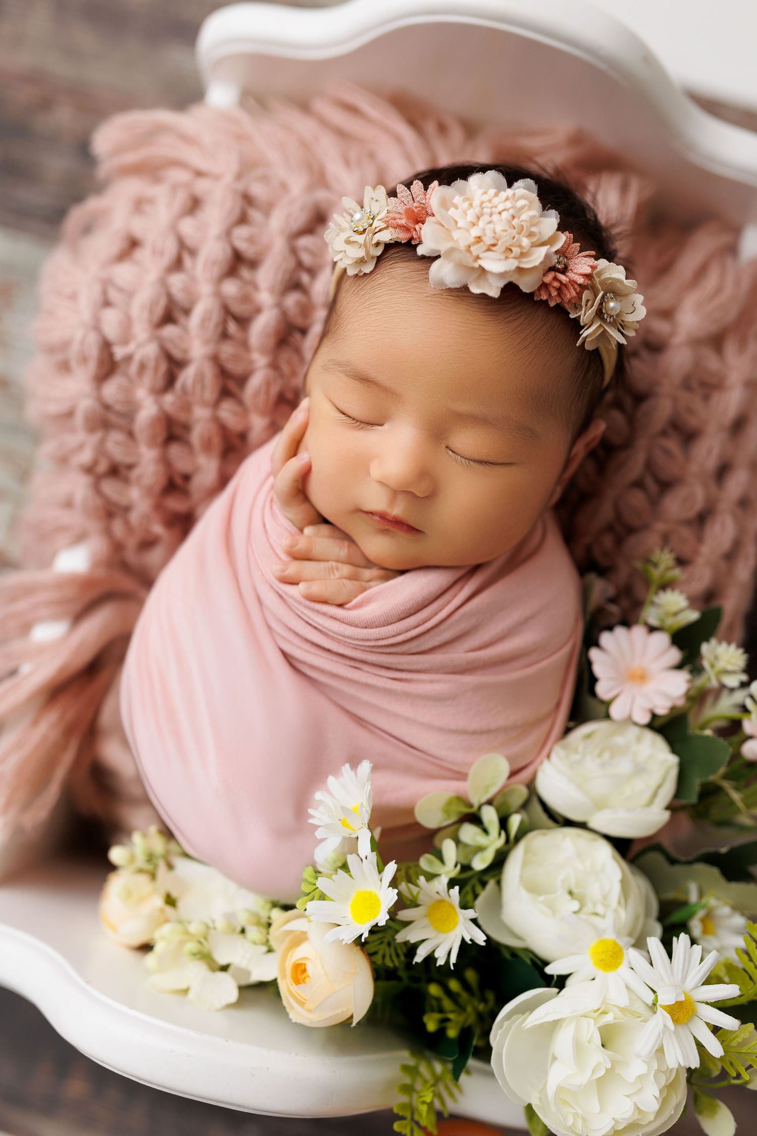 Sleeping newborn wrapped in soft pink fabric surrounded by delicate white daisies and roses in a dreamy portrait setting.