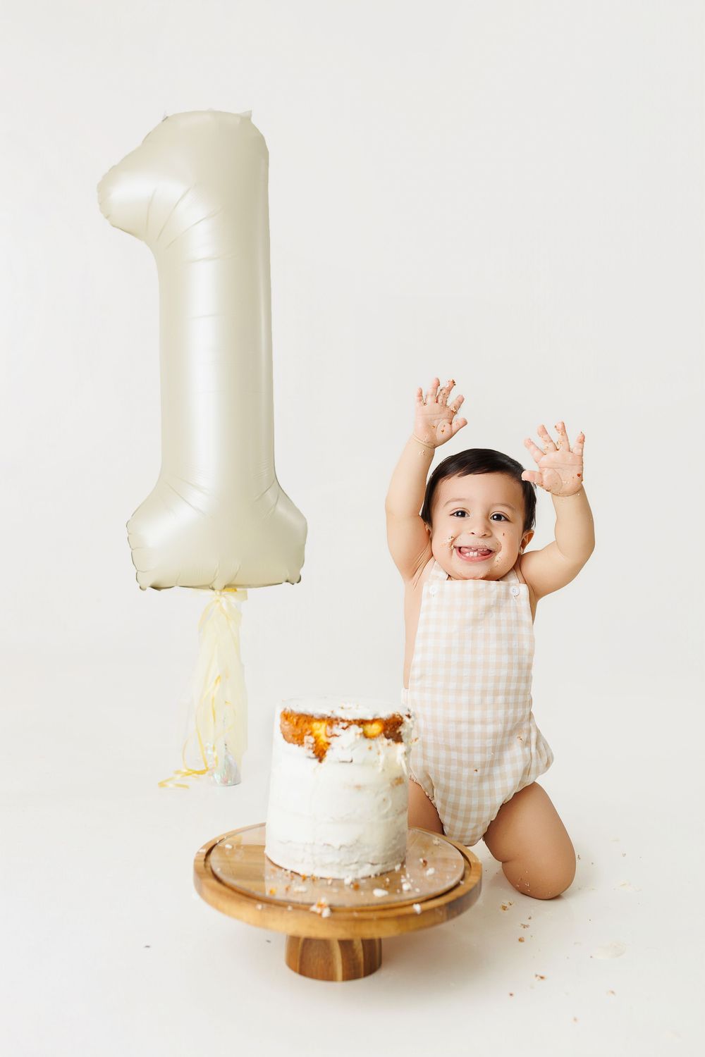 First birthday cake smash photo session with white number one balloon and gingham outfit on wooden backdrop.