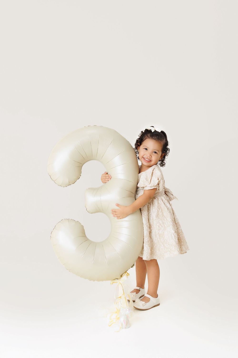 Sequence of four photos showing a child in white dress playing with a large cream-colored number three decoration.
