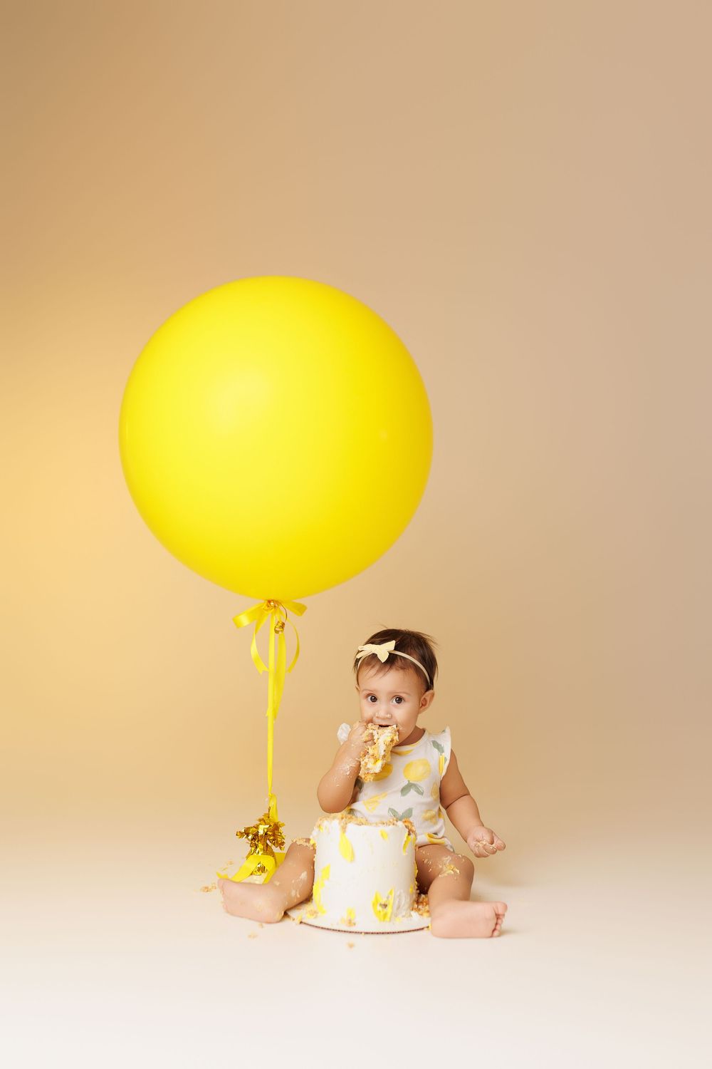 Baby in white outfit plays with yellow balloon on light beige background in progression photo series.