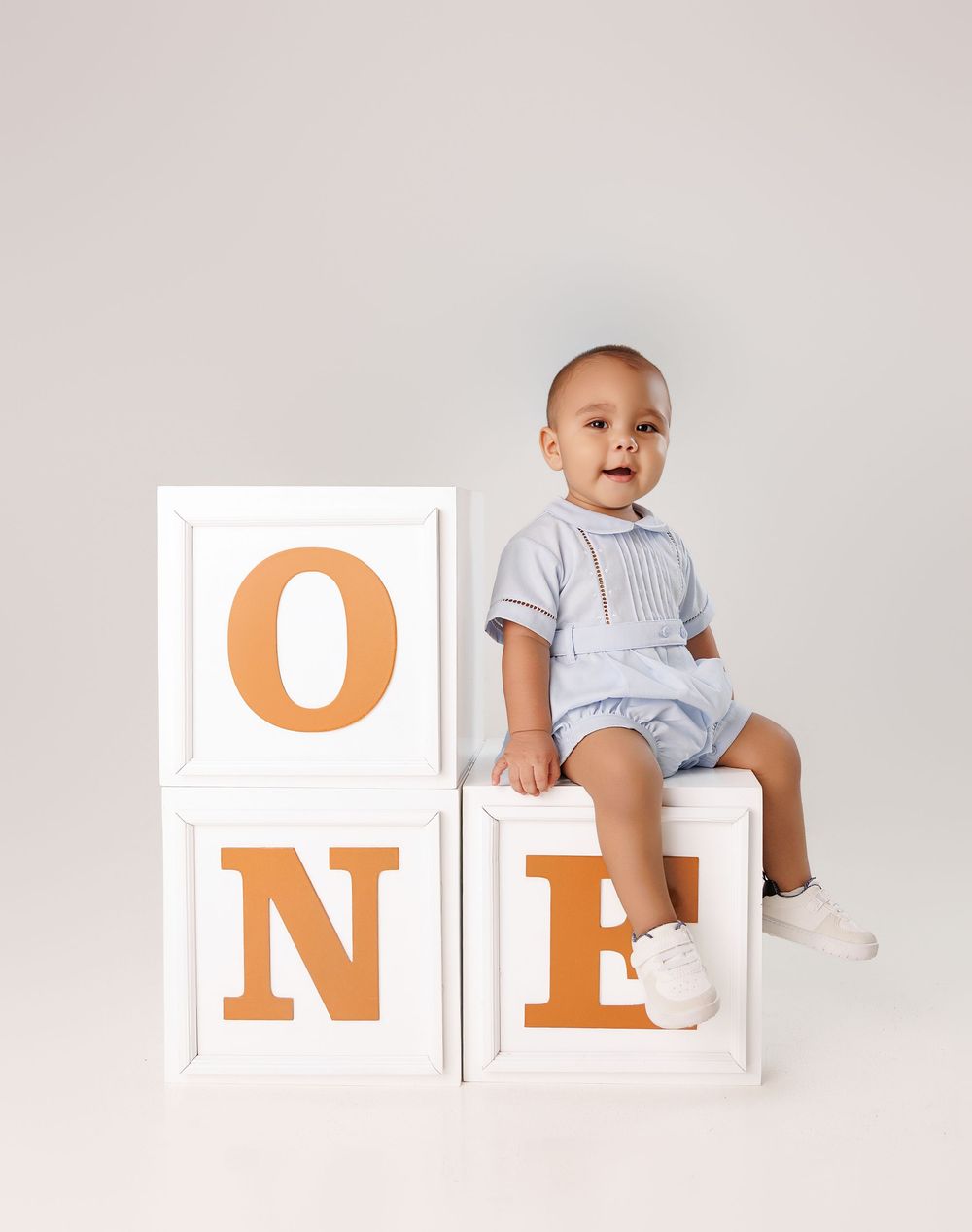 Baby wearing white onesie sits among orange letter blocks spelling ONE against white background.