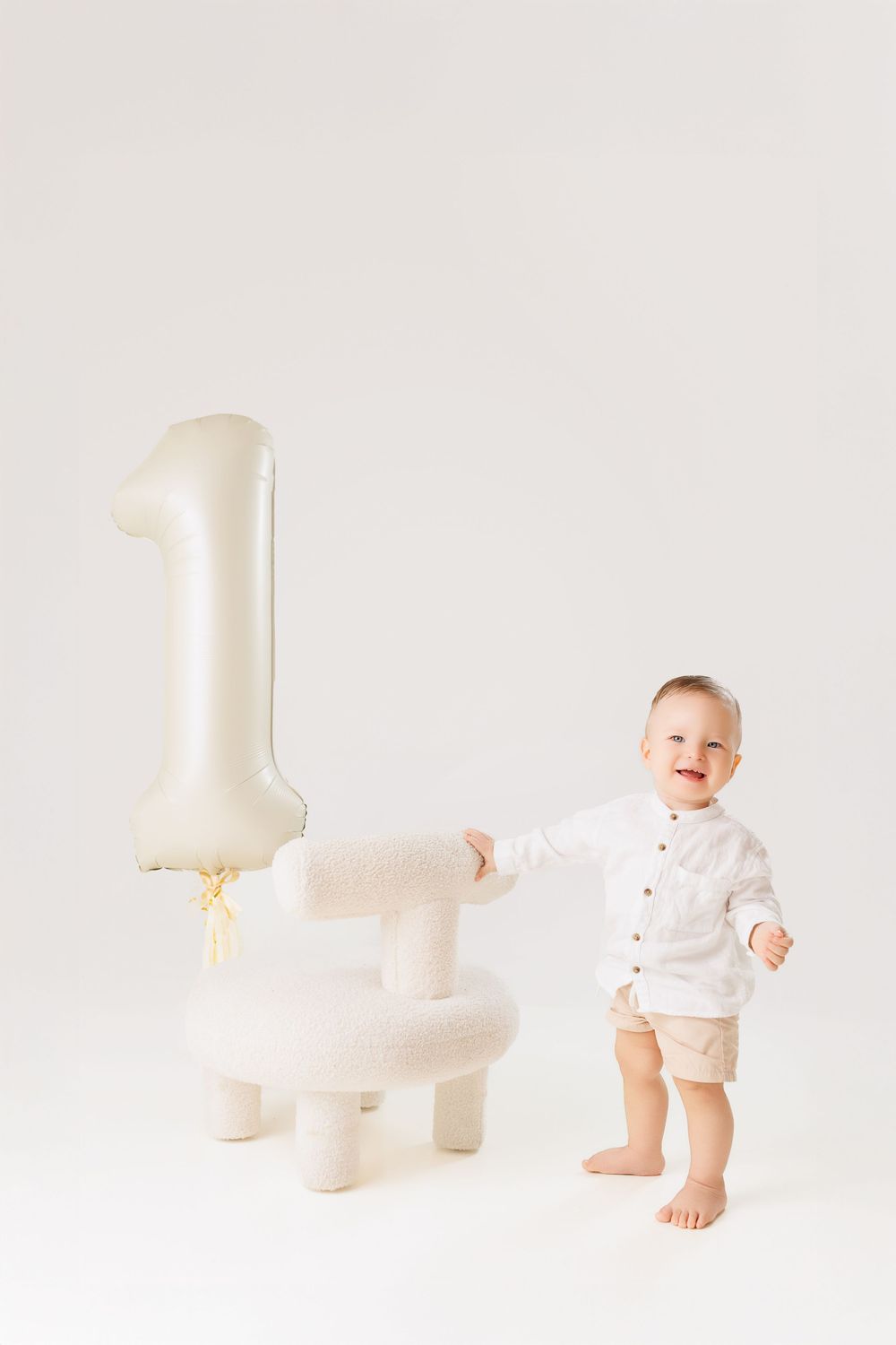 Baby in white onesie sits on floor with minimalist white toys and decor in a bright clean setting.