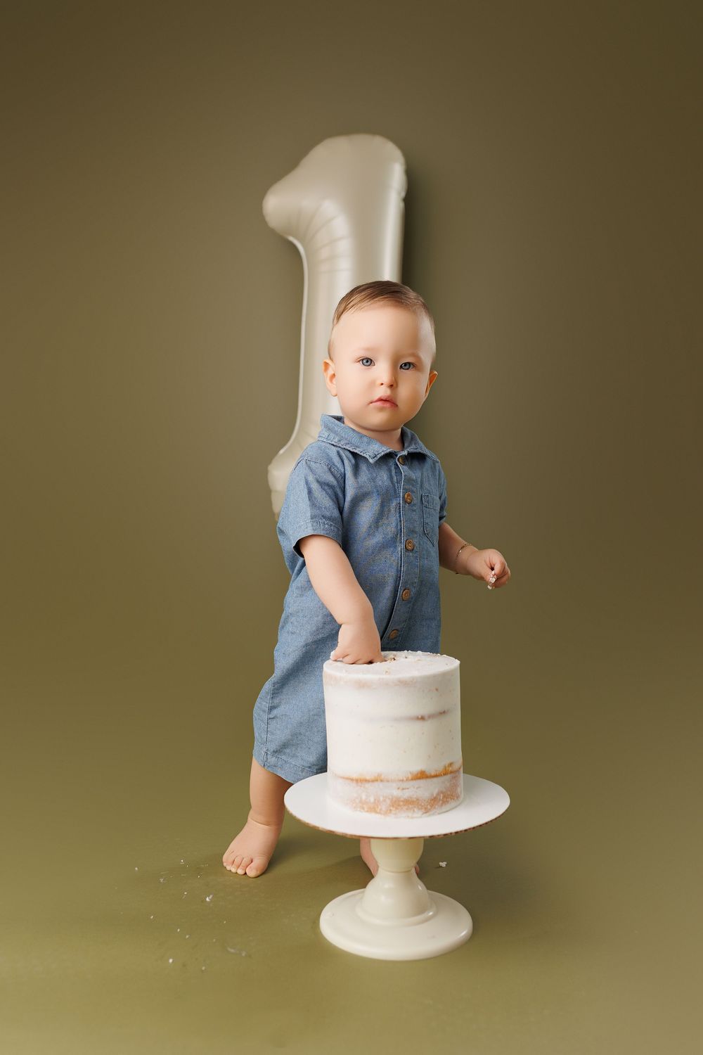 First birthday celebration with a toddler in denim outfit standing next to white cake on pedestal and large number one backdrop.