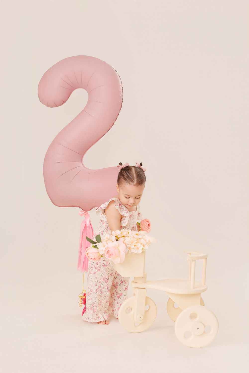 Baby in floral dress plays with pink toys and decor in soft pastel studio setting.