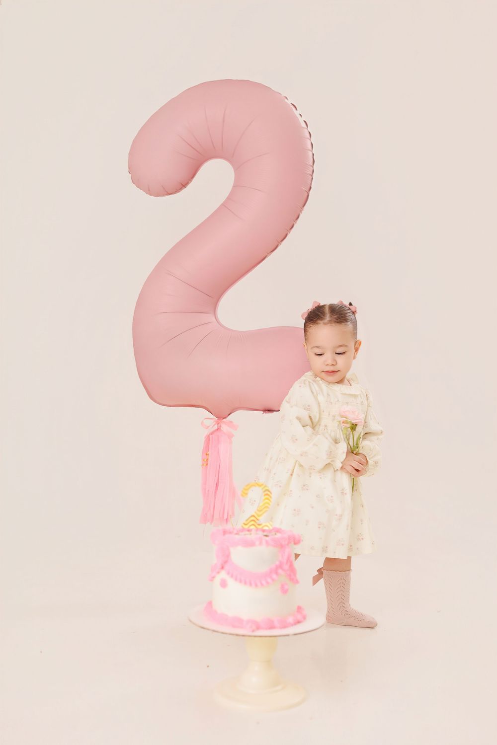 Toddler in white dress sits next to pink birthday cake with large number 2 balloon for second birthday photoshoot.