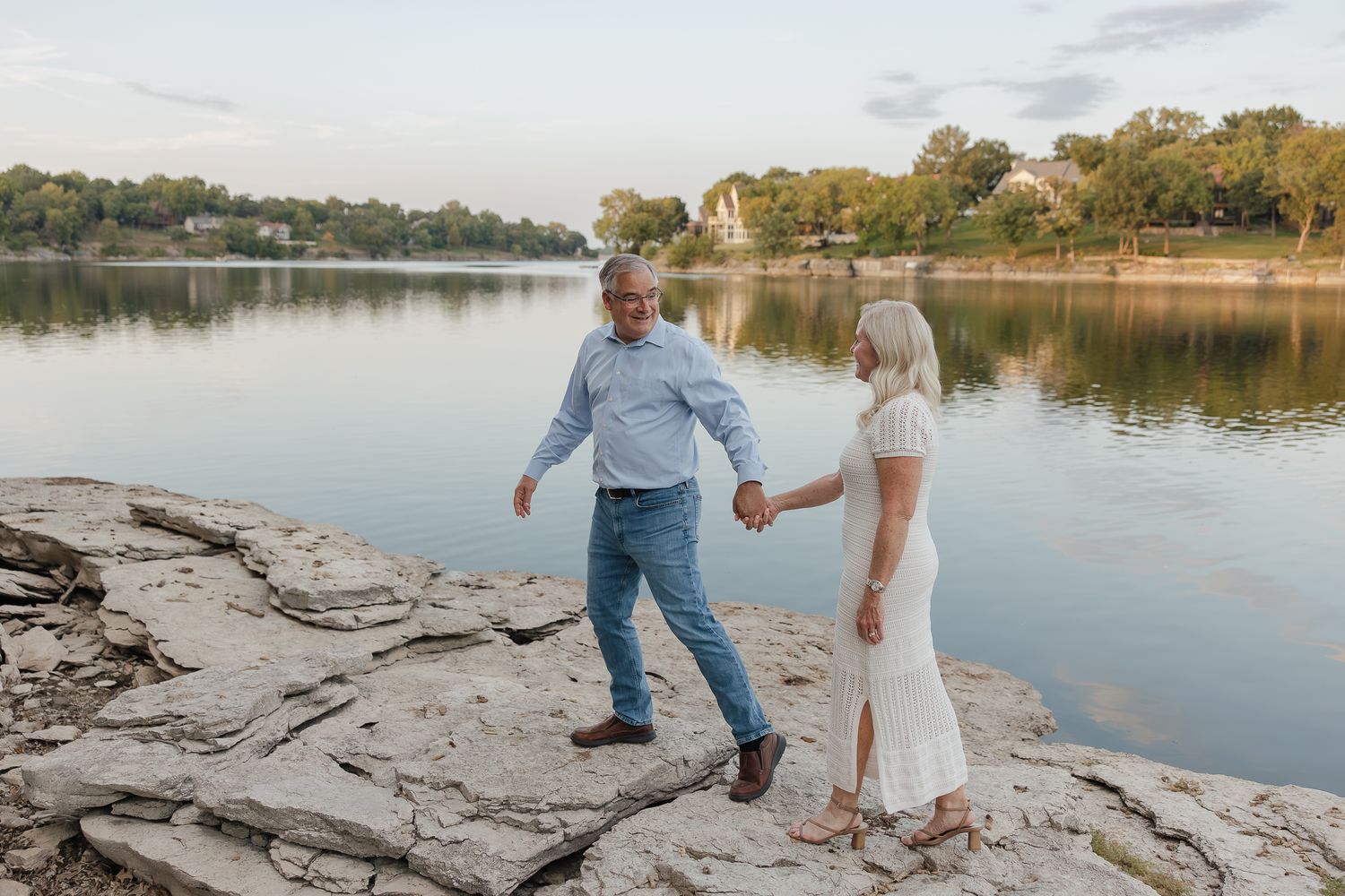 husband and wife holding hands at sunset walking along rock by lee summit lake