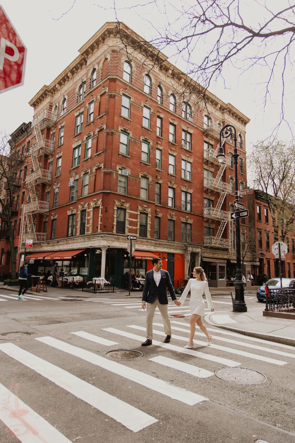Two people cross a zebra crossing in front of a historic red brick apartment building in an urban neighborhood.
