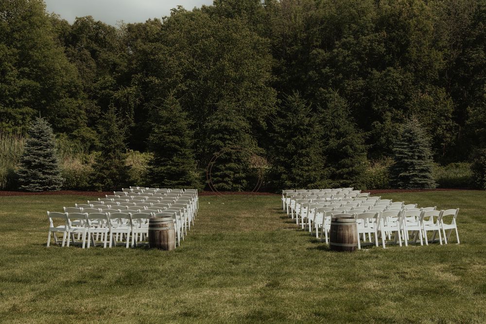 White wedding chairs arranged in curved rows on a green lawn surrounded by tall trees for an outdoor ceremony.