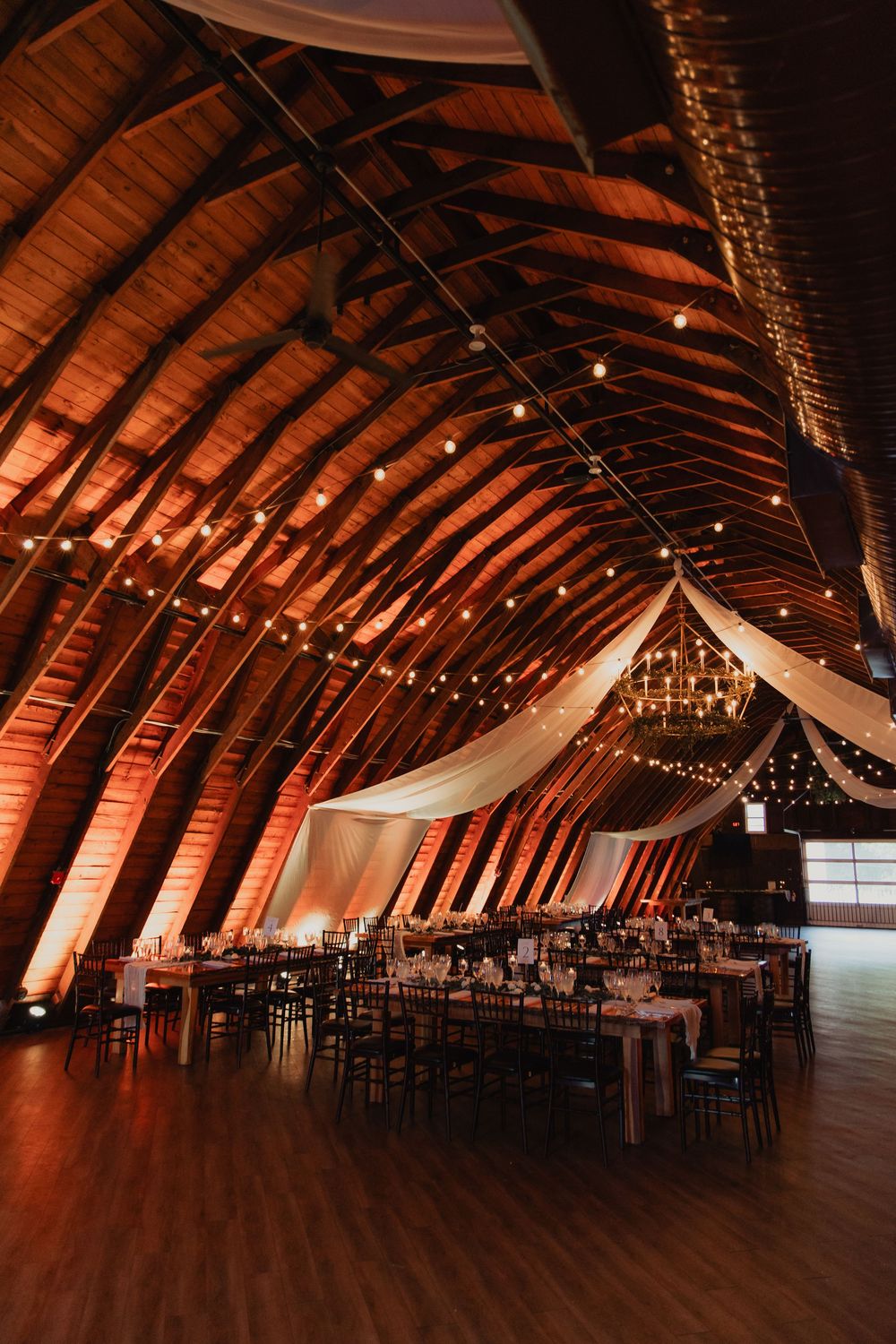 Rustic barn wedding venue interior with exposed wooden beams, string lights and white fabric draping across vaulted ceiling.