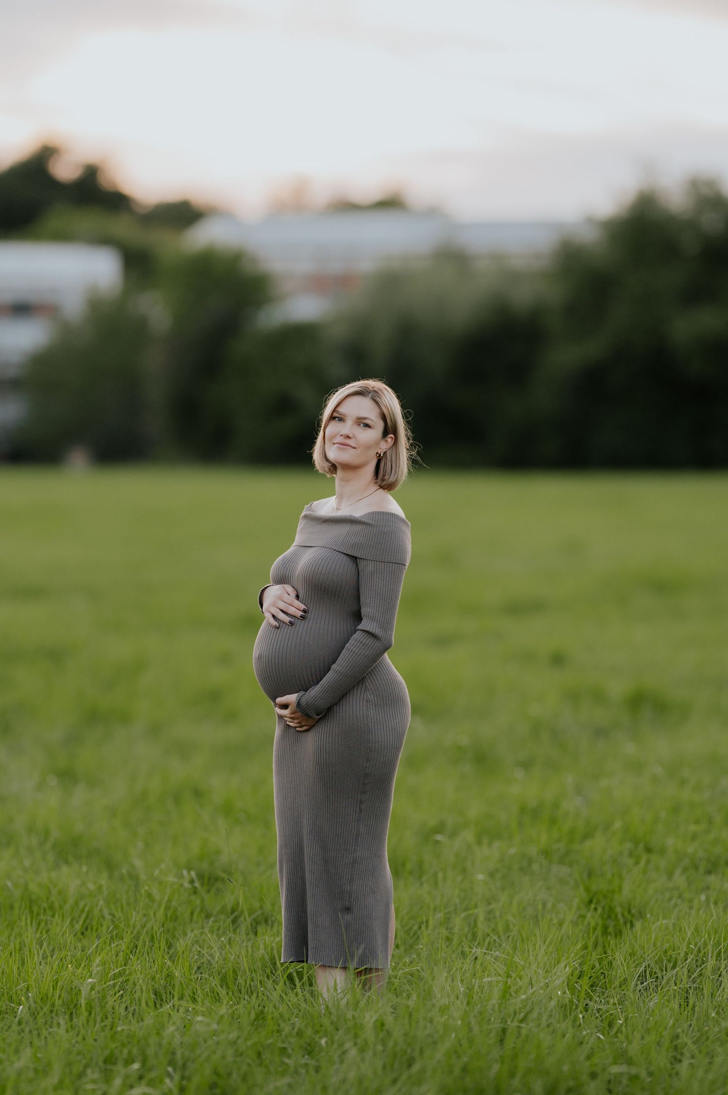 Eine schwangere Frau in einem grauen Kleid steht während eines Schwangerschaftsfotoshootings bei Sonnenuntergang auf einer Wiese.