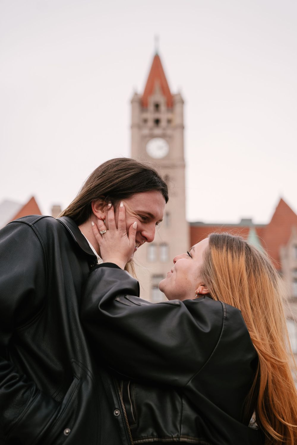 Romantic couple in black leather jackets sharing a tender moment with a clock tower in the background.