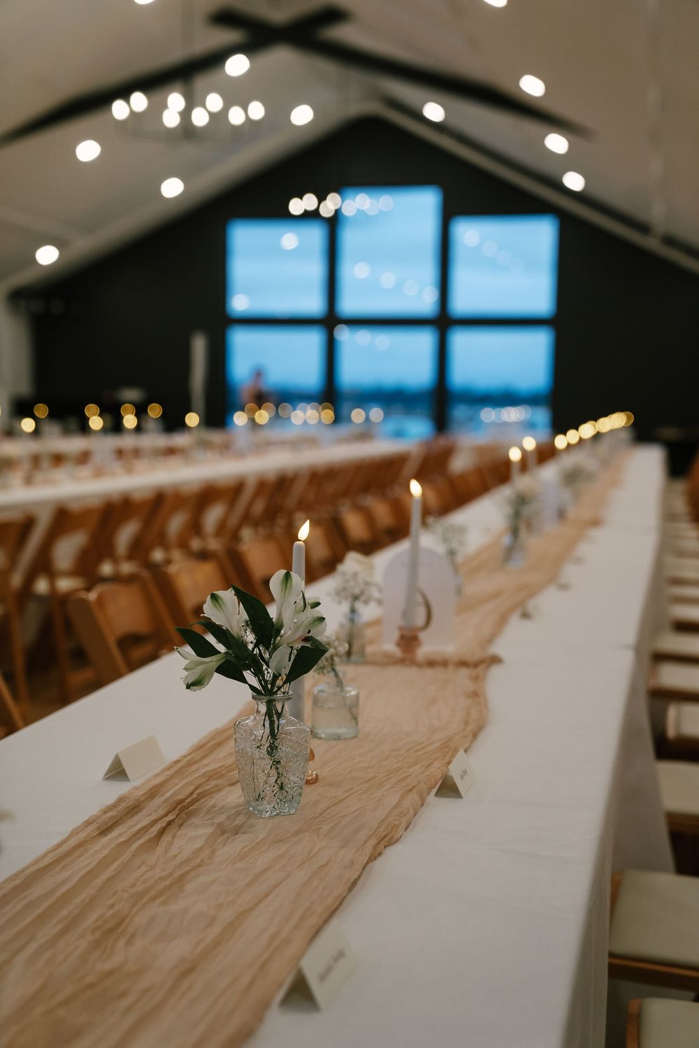 Elegant wedding reception hall with long white tables, burlap runners, candles, and floral arrangements under warm string lights.