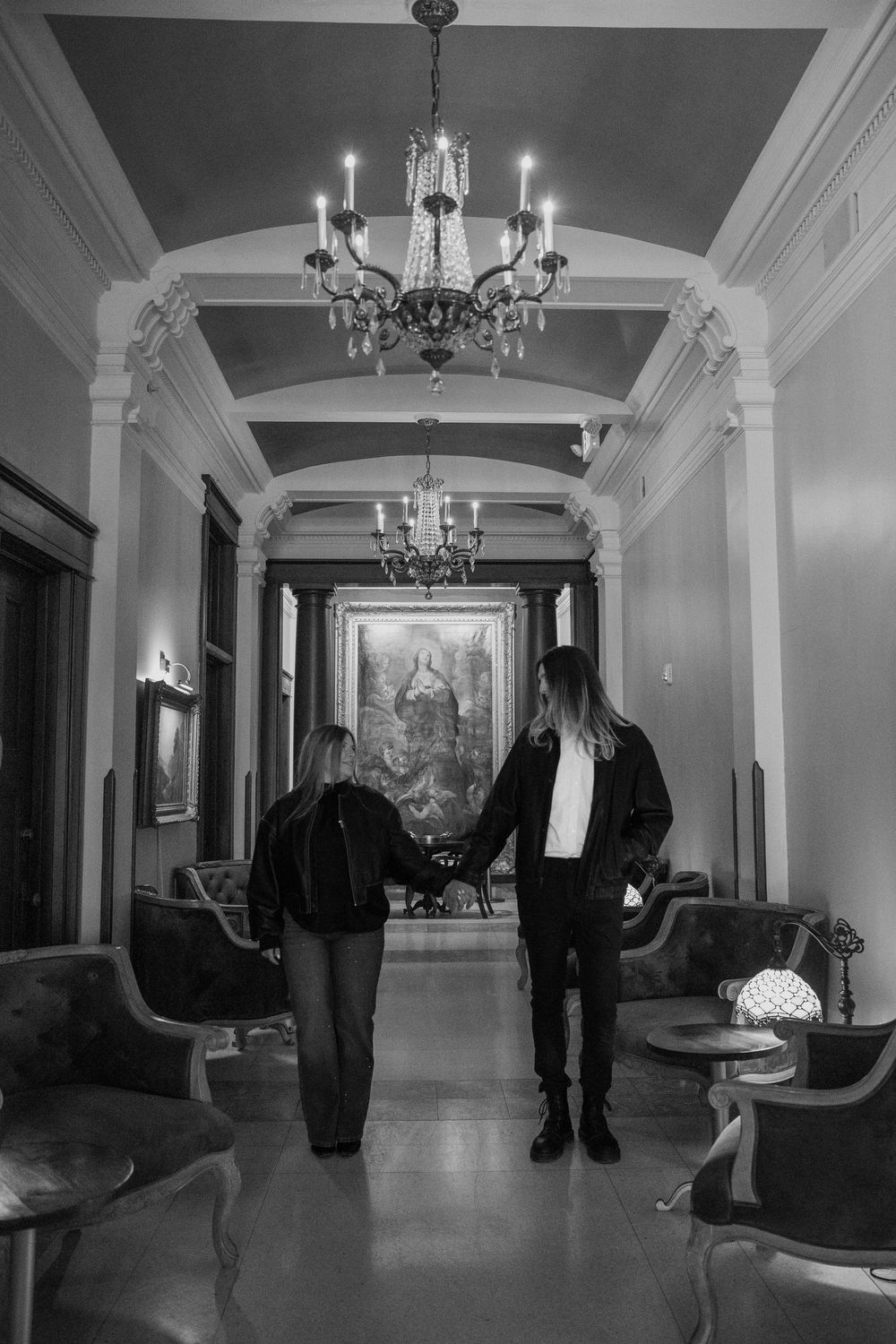 Two people hold hands walking through an elegant black and white hallway with chandeliers and classic artwork.