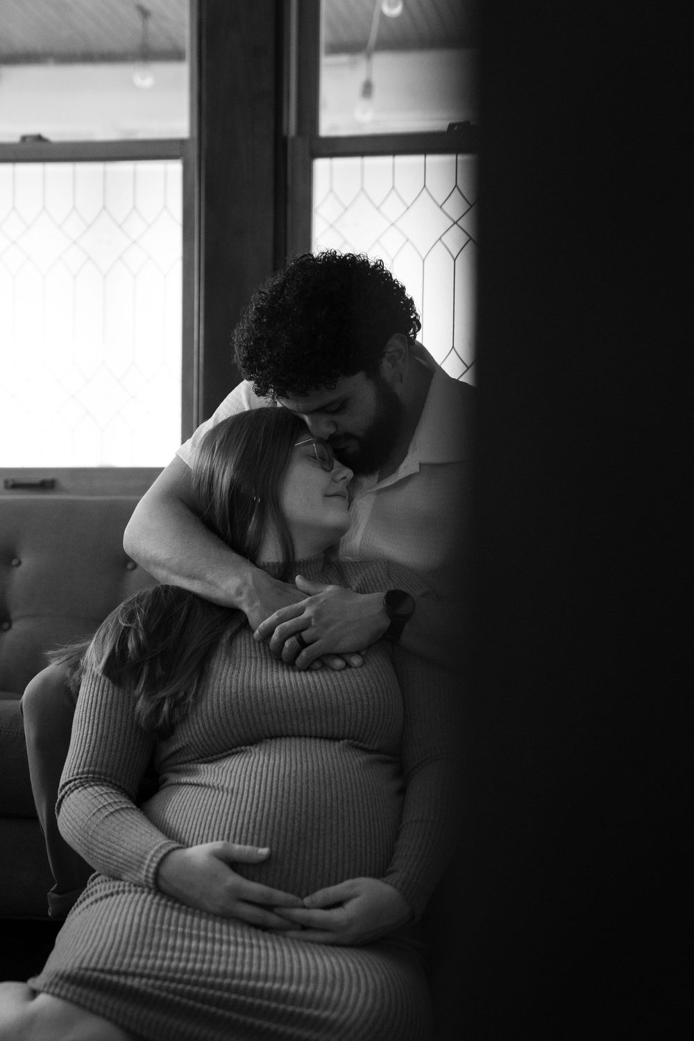 Black and white photo of a couple sharing a tender moment, man kissing pregnant woman on forehead while she cradles her baby bump.