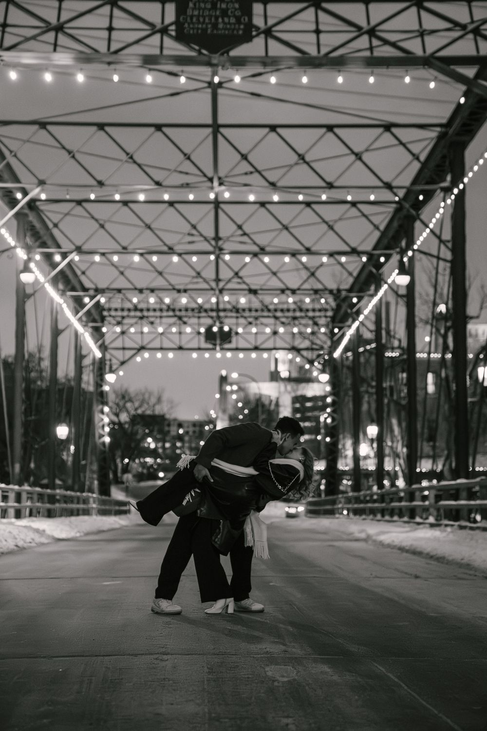 Couple embracing on a bridge at night with string lights overhead in black and white photo.